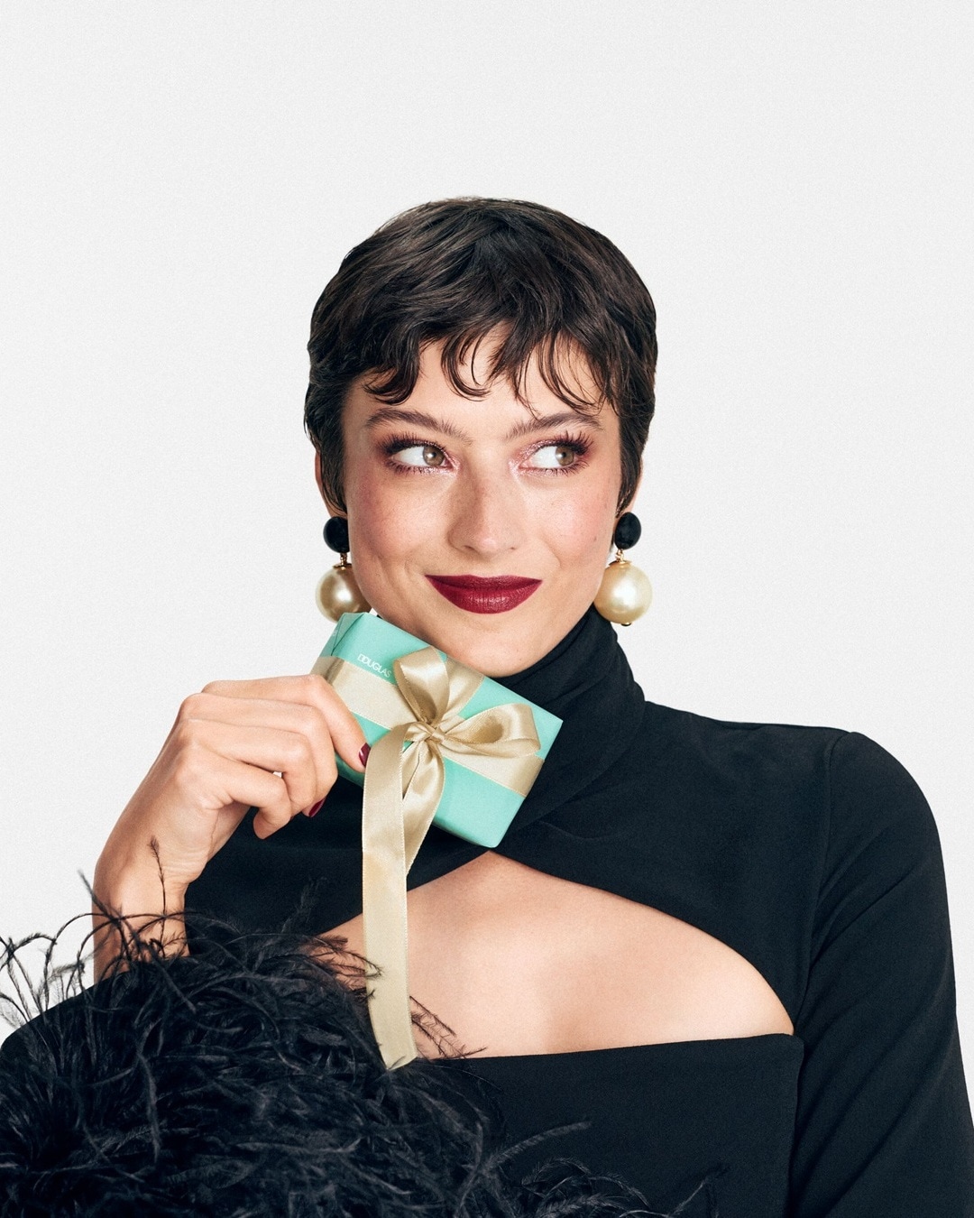 A képen egy rövid hajú, fekete ruhát viselő nő látható, aki egy világoskék Douglas ajándékutalványt tart a kezében, arany masnival átkötve.