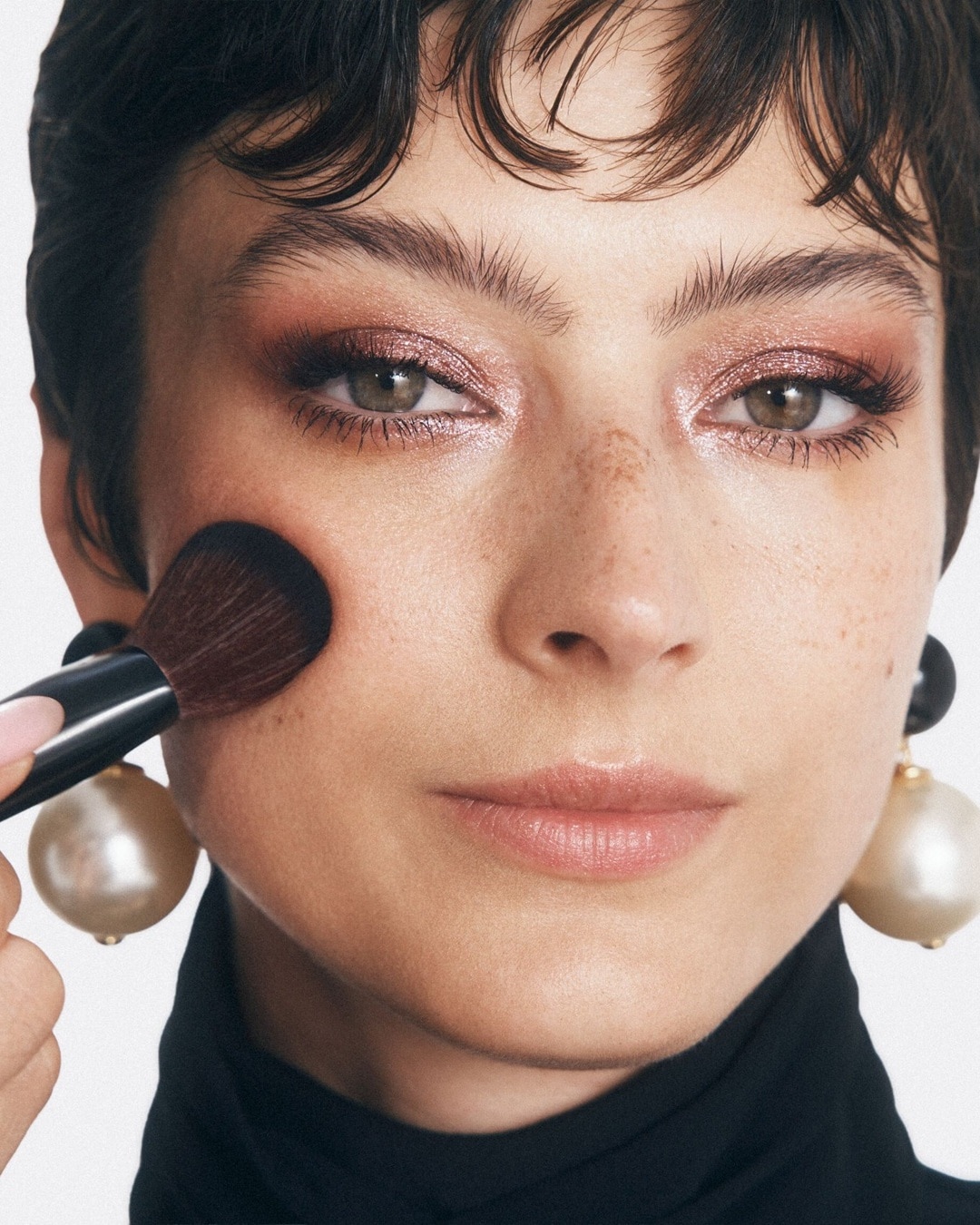 A képen egy nő látható, amint éppen sminkelik: egy ecsettel pirosítót visznek fel az arcára, a szeme rózsaszín csillogó szemhéjfestékkel van kiemelve, és nagy gyöngy fülbevalót visel.