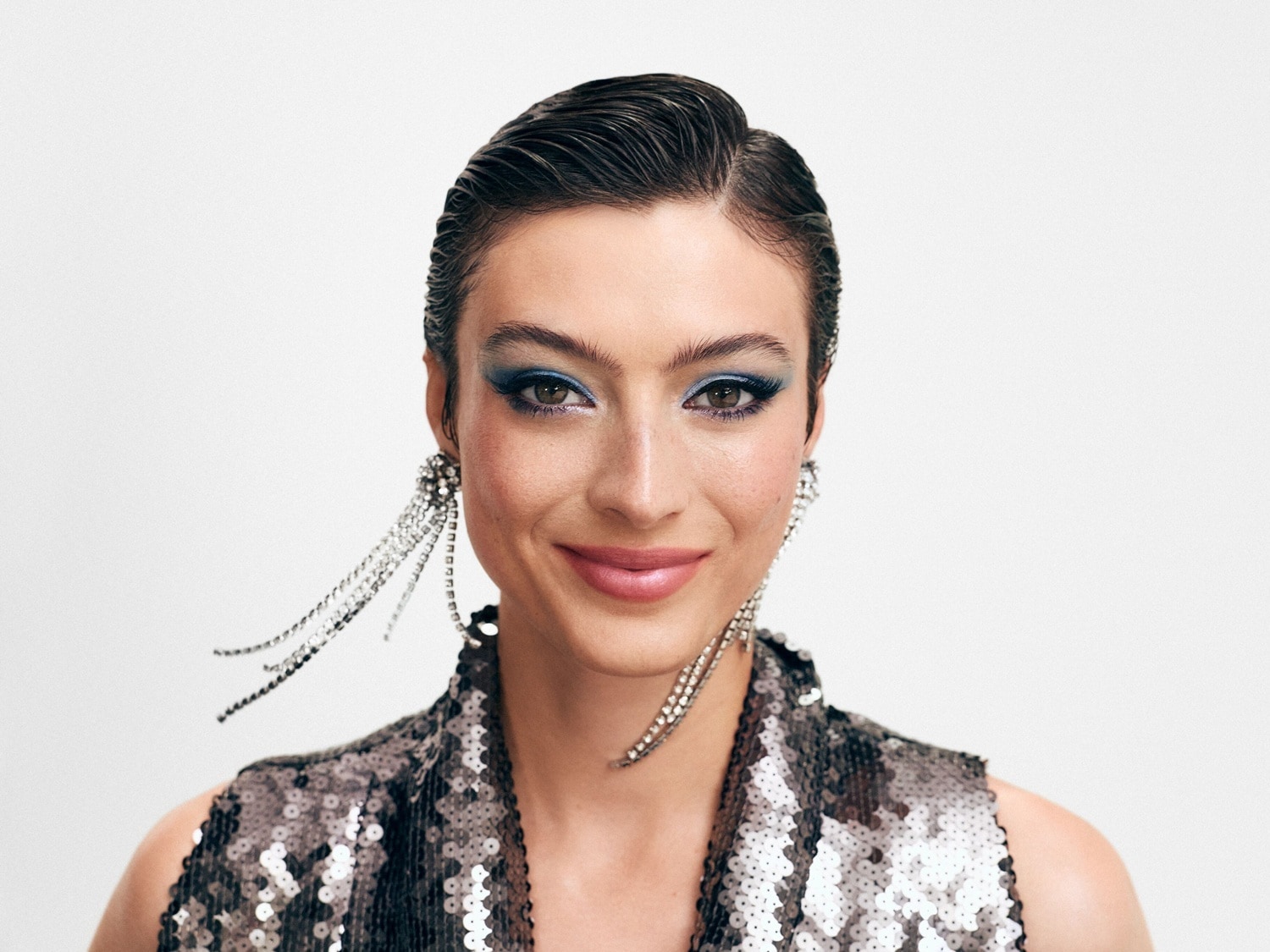 Moters portretas su blizgančiu makiažu, mėlynais akių šešėliais ir sidabriniais auskarais su kutais, vilkinčios blizgučiais dekoruotą viršutinę dalį.