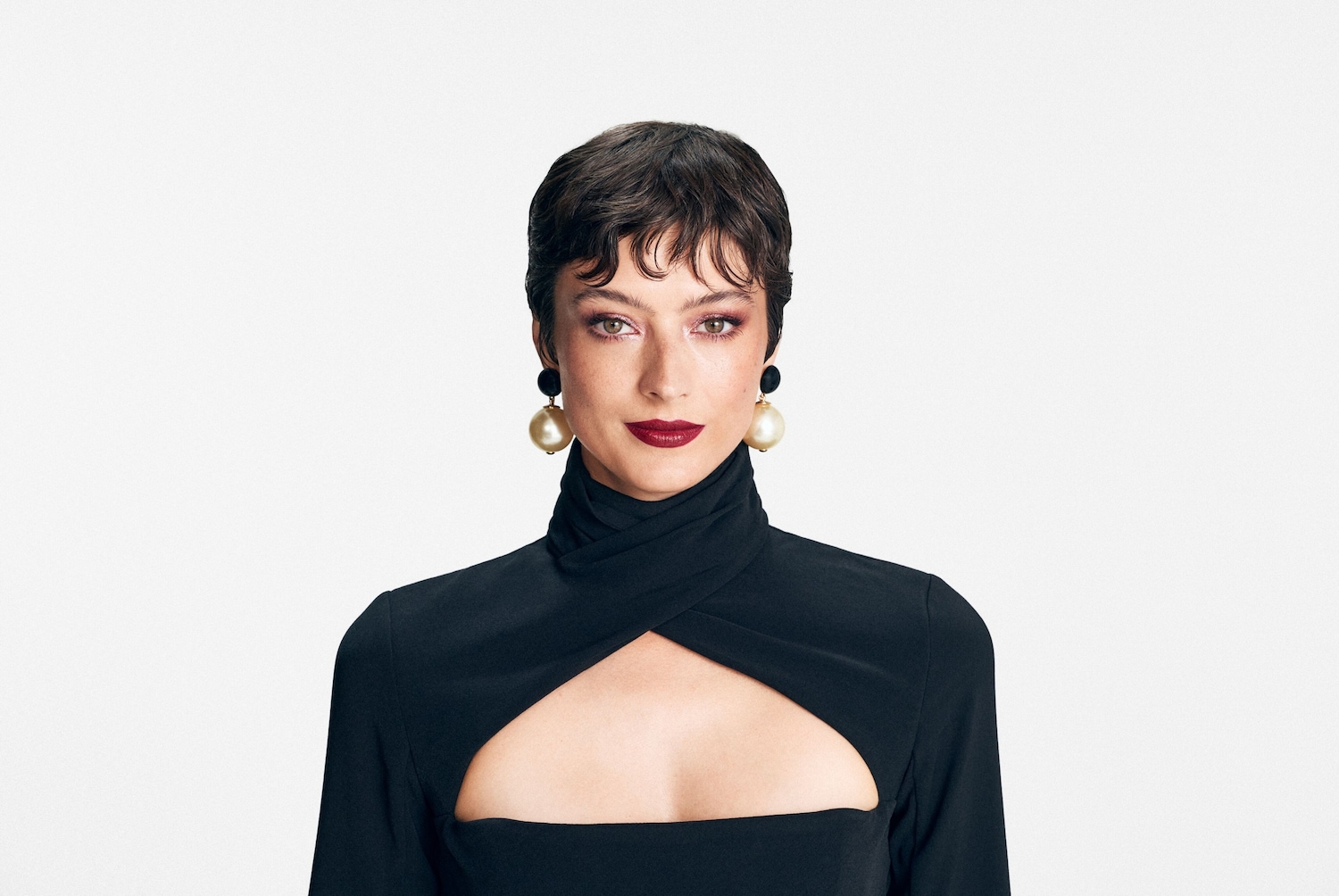 A képen egy rövid, barna hajú, világos bőrű nő látható, aki fekete, kivágott ruhát visel, és nagy gyöngy fülbevalót hord.