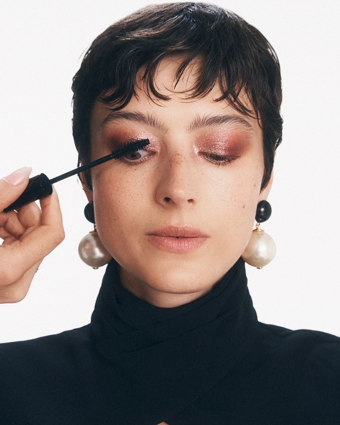 A képen egy nő látható, aki éppen fekete szempillaspirált visz fel a felső szempilláira, a szeme vöröses-barna csillogó szemhéjfestékkel van festve, és nagy gyöngy fülbevalót visel.