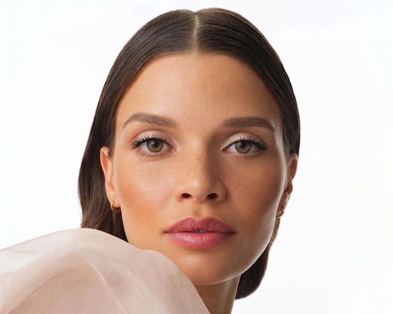 Primo piano di una donna con occhi verdi, ombretto scintillante color champagne e labbra rosa chiaro, che indossa un top rosa pallido.