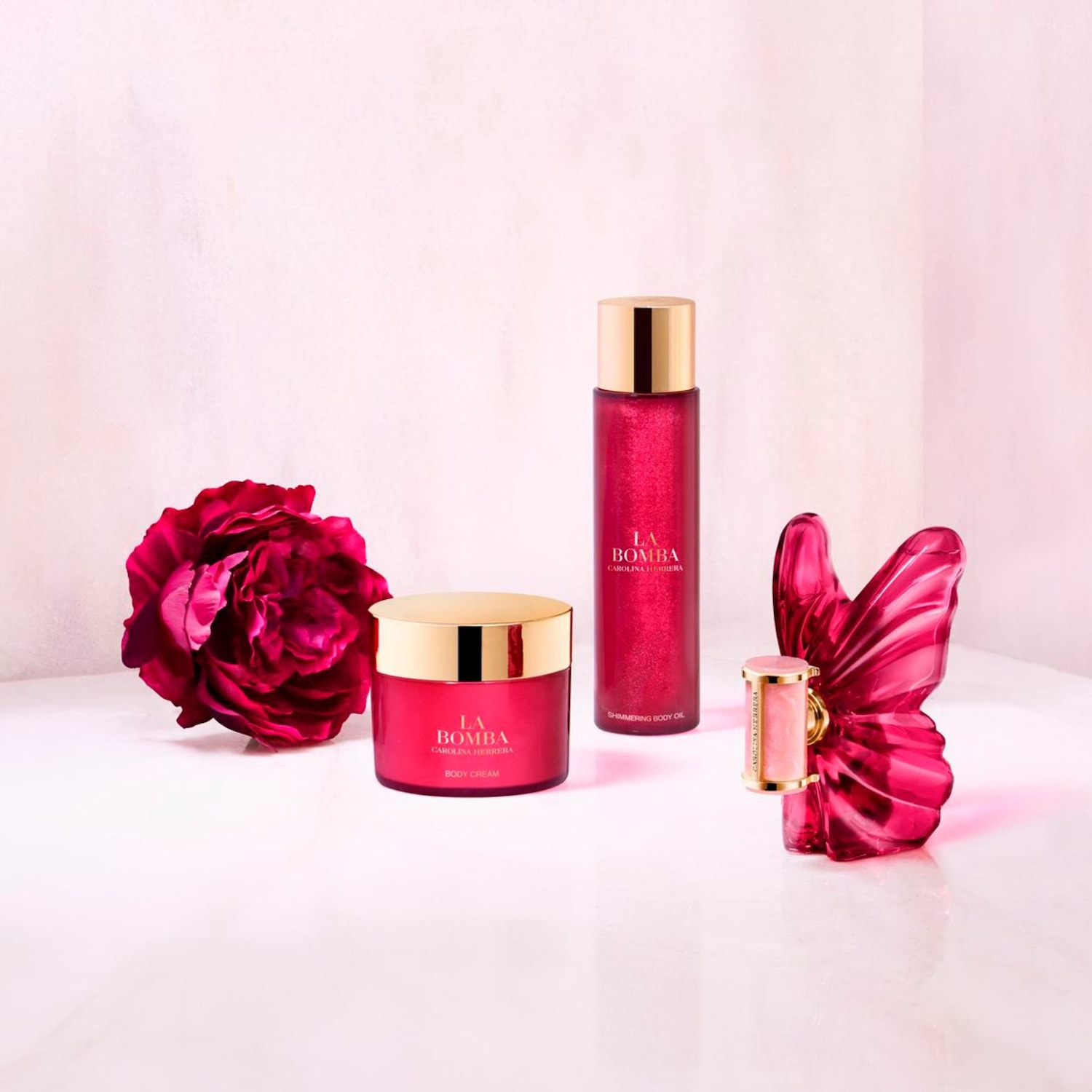 Zářivá sada péče o tělo La Bomba Carolina Herrera s tělovým krémem, třpytivým olejem a ozdobným motýlem, vše v odstínech růžové a zlaté.