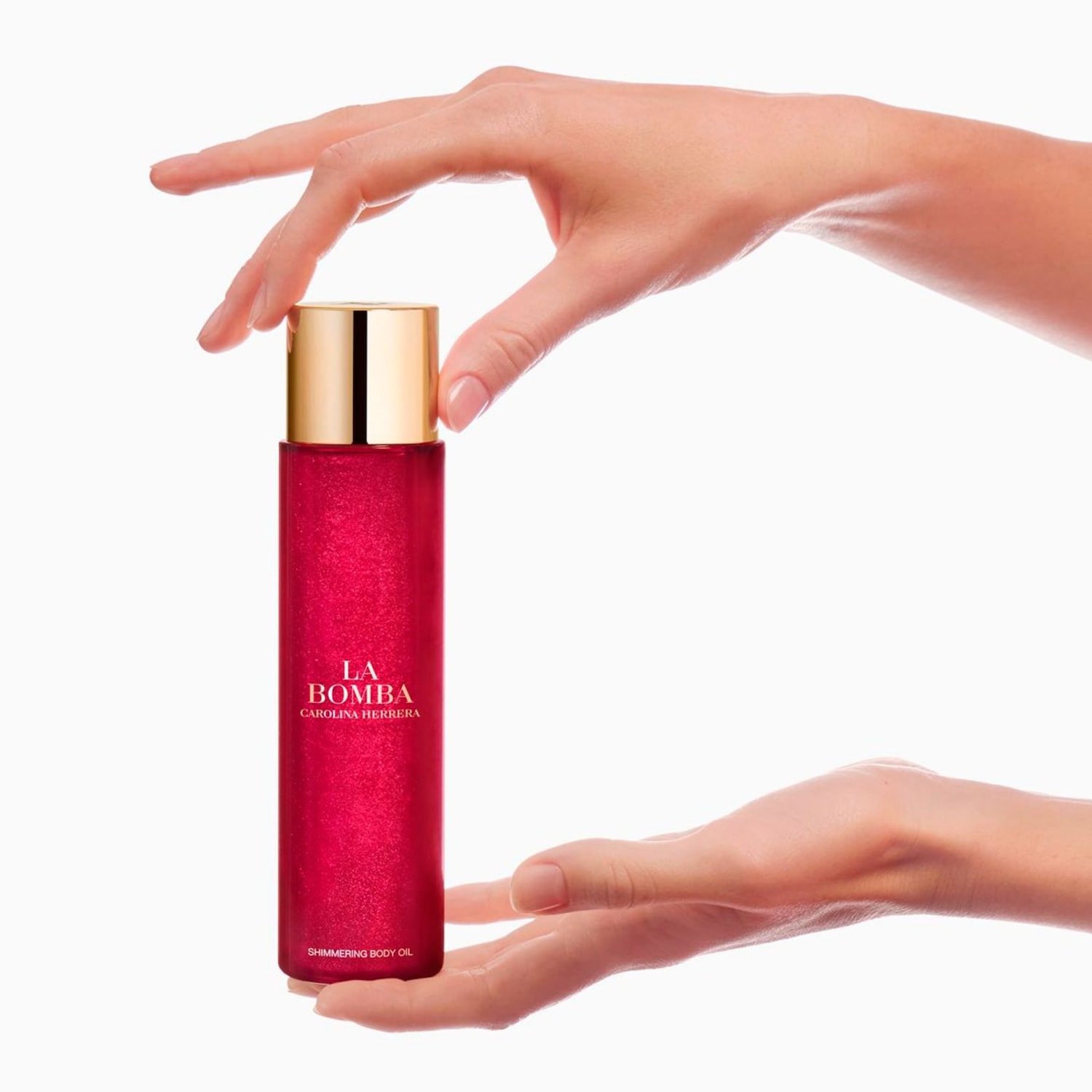 Carolina Herrera La Bomba třpytivý tělový olej v červené lahvičce se zlatým víčkem, držený v rukou.
