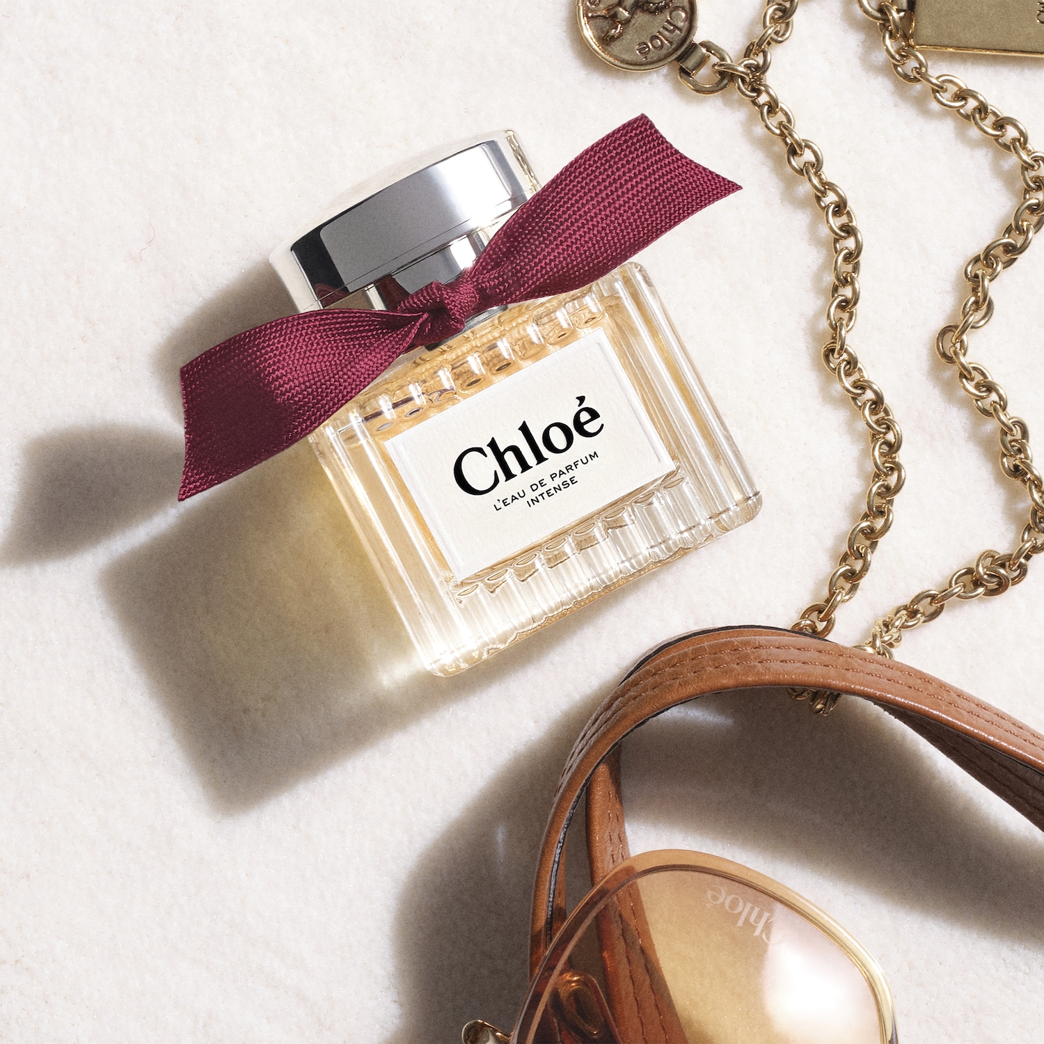 Butelka perfum Chloé L'Eau de Parfum Intense z bordową wstążką, leżąca obok złotego łańcuszka i okularów przeciwsłonecznych Chloé.