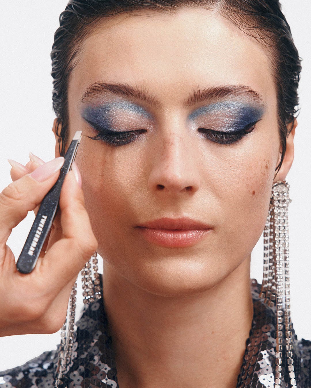 Moters veidas su ryškiu mėlynu akių makiažu, ranka laiko pincetą prie blakstienų, pabrėžiant grožio procedūrą.