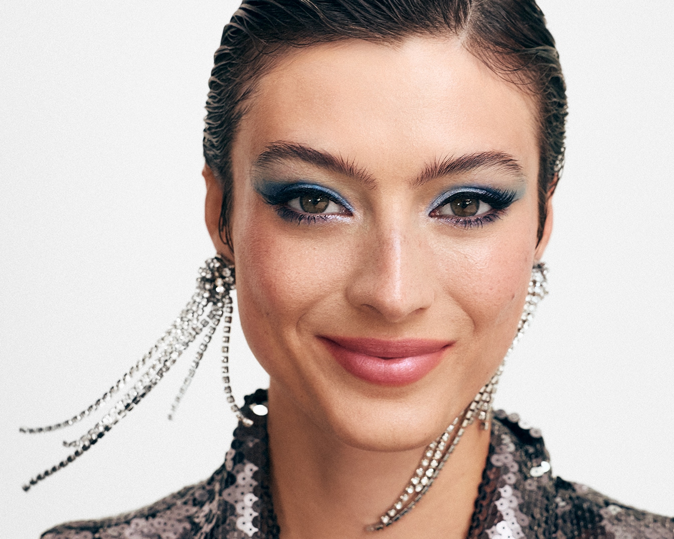 Gros plan d'une femme souriante avec un fard à paupières bleu scintillant et de longues boucles d'oreilles à chaînes argentées, portant une veste à paillettes.