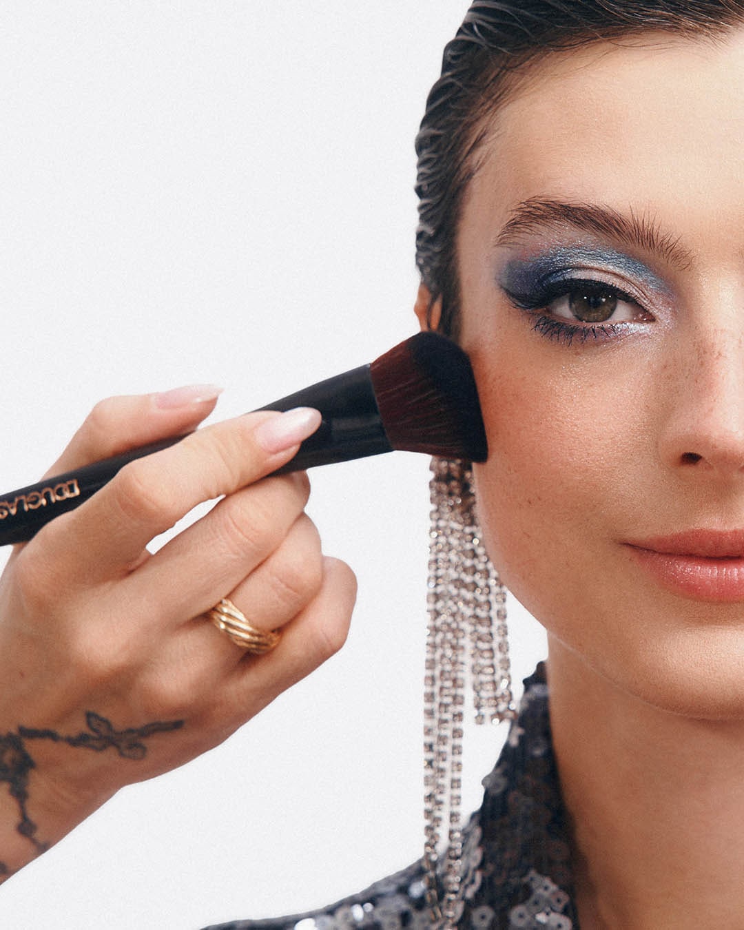 Šepetėliu tepamas skaistalas ant moters skruosto, ji dėvi blizgučiais dekoruotą viršutinę dalį ir ilgus auskarus su kutais, o akys padažytos mėlynais šešėliais.