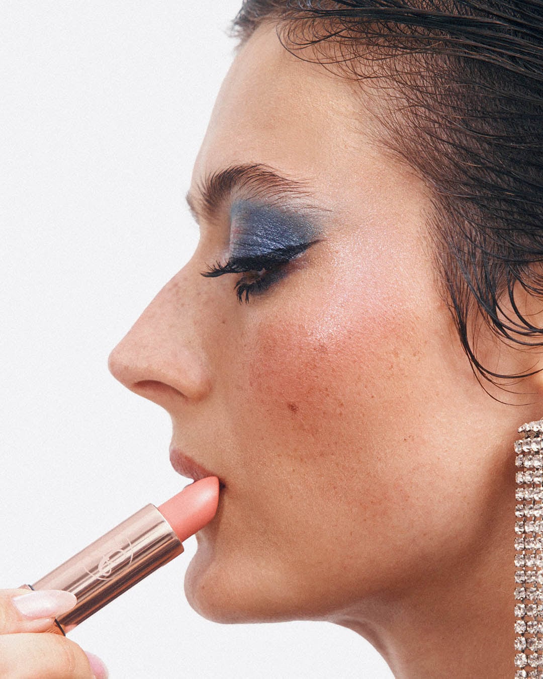 Šoninis moters portretas su mėlynais akių šešėliais ir rožiniu lūpdažiu, tepamu auksiniu aplikatoriumi, papuoštas blizgančiu auskaru.