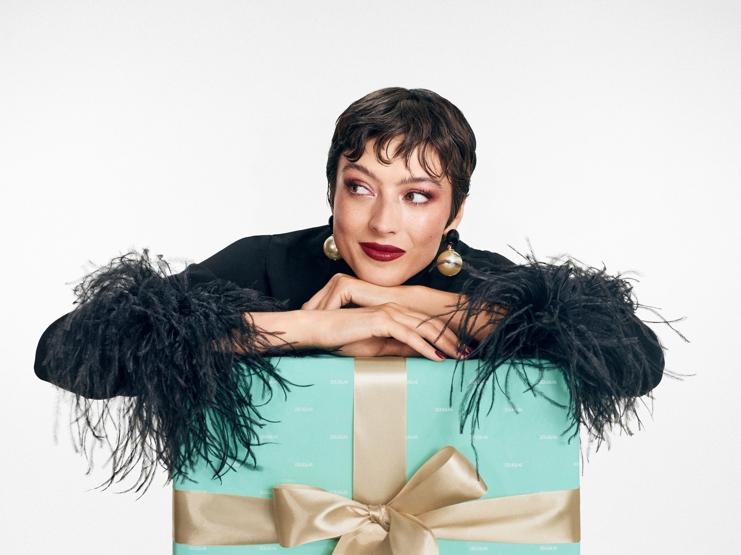 Elegantiška moteris su juodu kostiumėliu, papuoštu plunksnomis, remiasi į dovanų dėžutę, perrištą auksiniu kaspinu, žvelgdama į šoną.