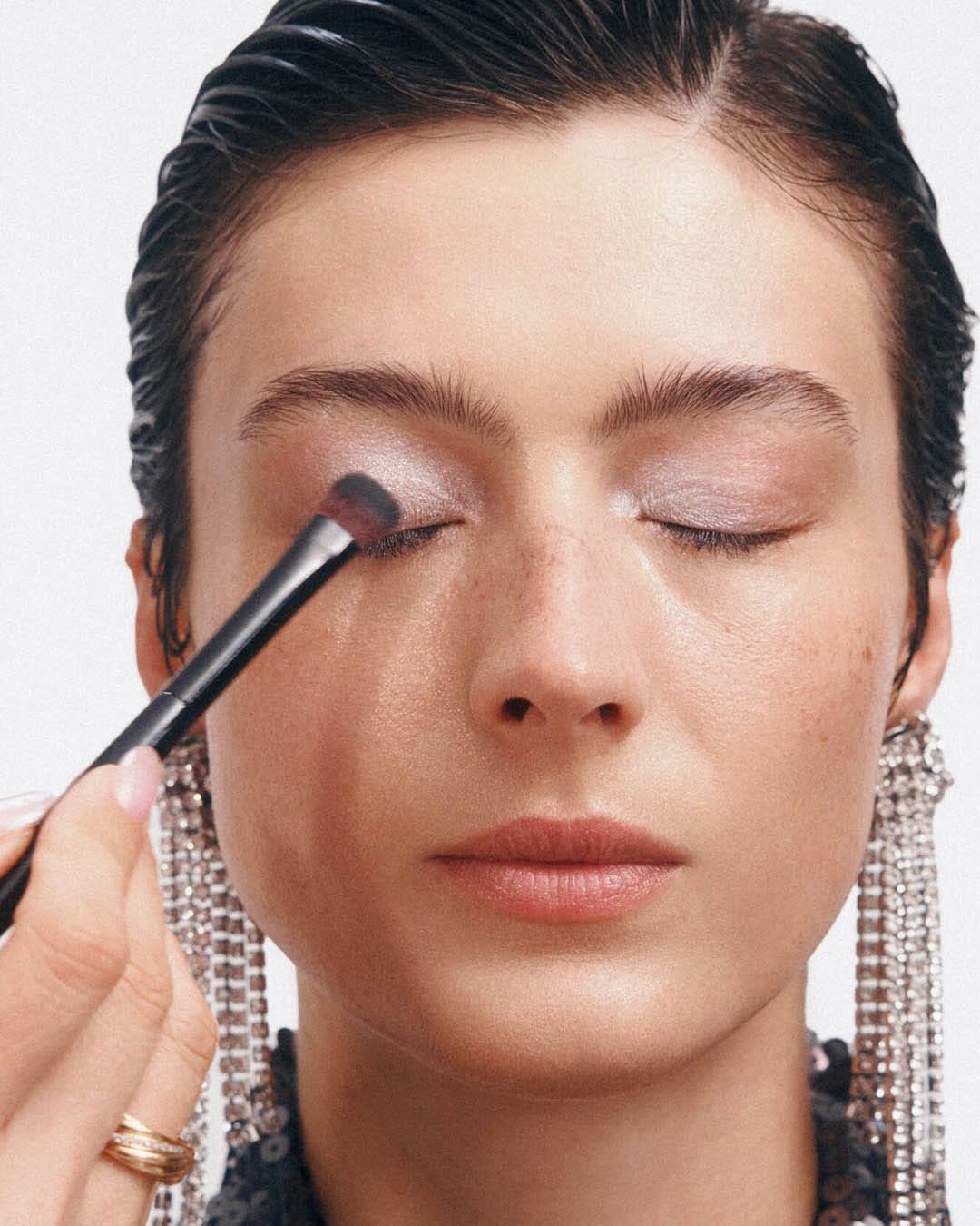 Šepetėliu tepami sidabriniai akių šešėliai ant užmerktų moters akių, kuri dėvi blizgančius auskarus.