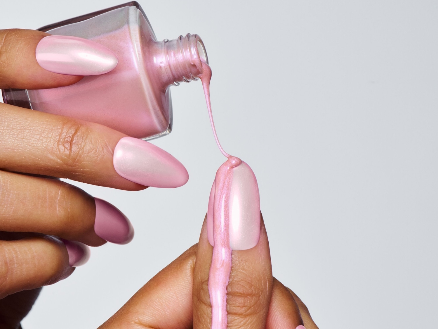 Eine Hand hält eine Flasche rosa Nagellack, der auf einen lackierten Fingernagel gegossen wird und so einen schimmernden, glänzenden Look erzeugt.