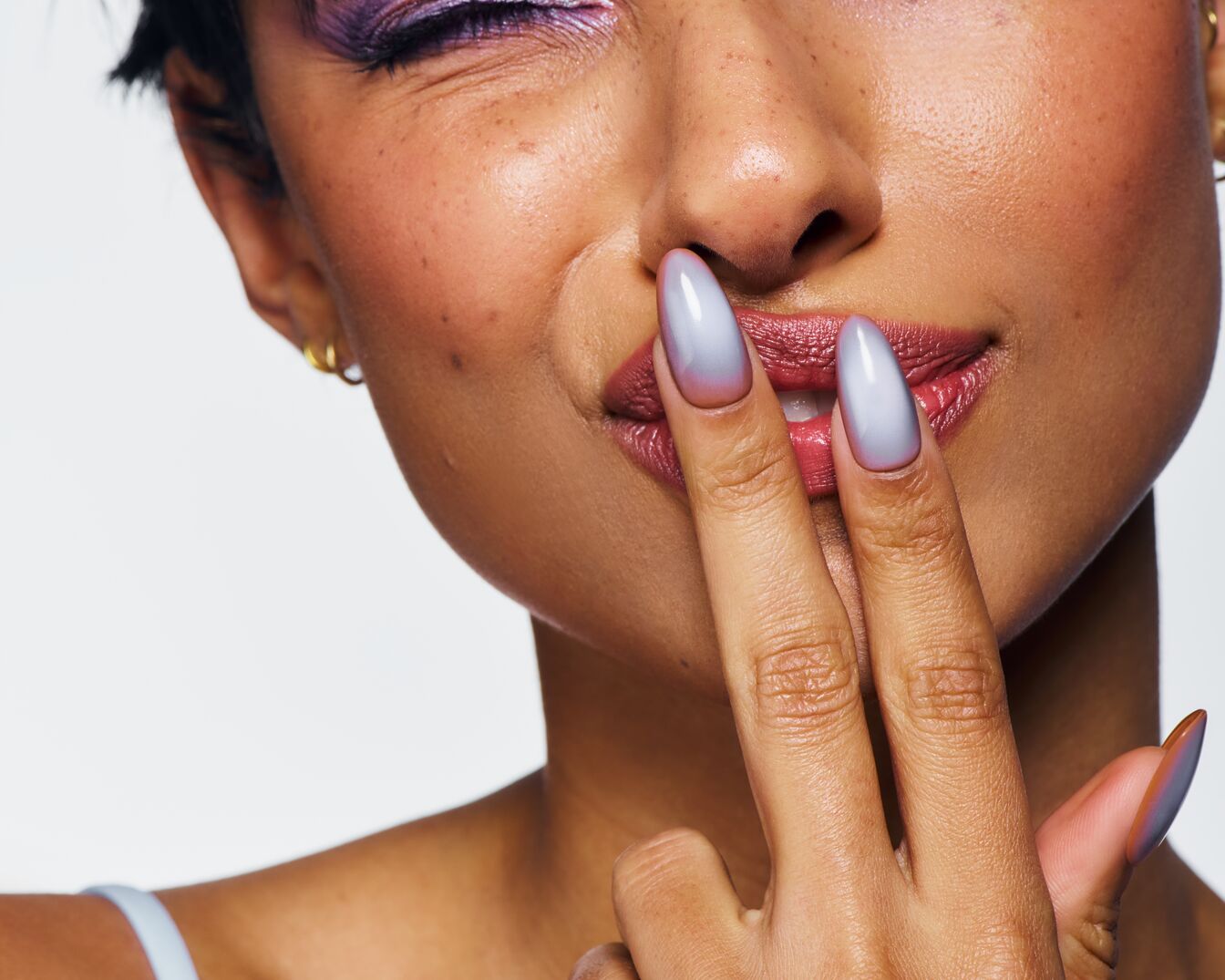 Nahaufnahme einer Frau mit lila Lidschatten und Ombre-Nägeln, die ihre Finger an die Lippen hält.