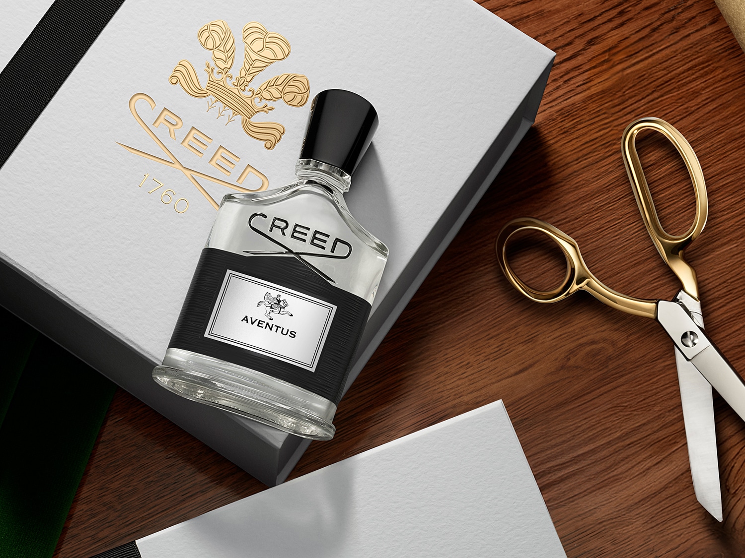 Auf einem Holztisch liegt eine Geschenkbox mit goldener Creed-Aufschrift, daneben eine Flasche Creed Aventus mit schwarzem Verschluss und goldene Schere.