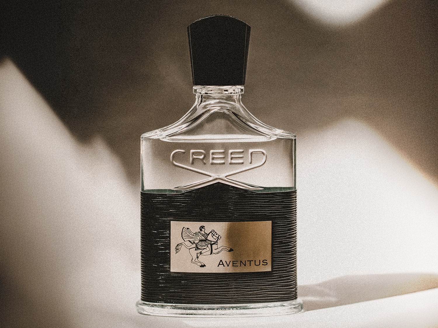Nahaufnahme eines Creed Aventus Parfümflakons mit klarem Glas, schwarzem Verschluss und einem goldenen Etikett, das einen Reiter zeigt.
