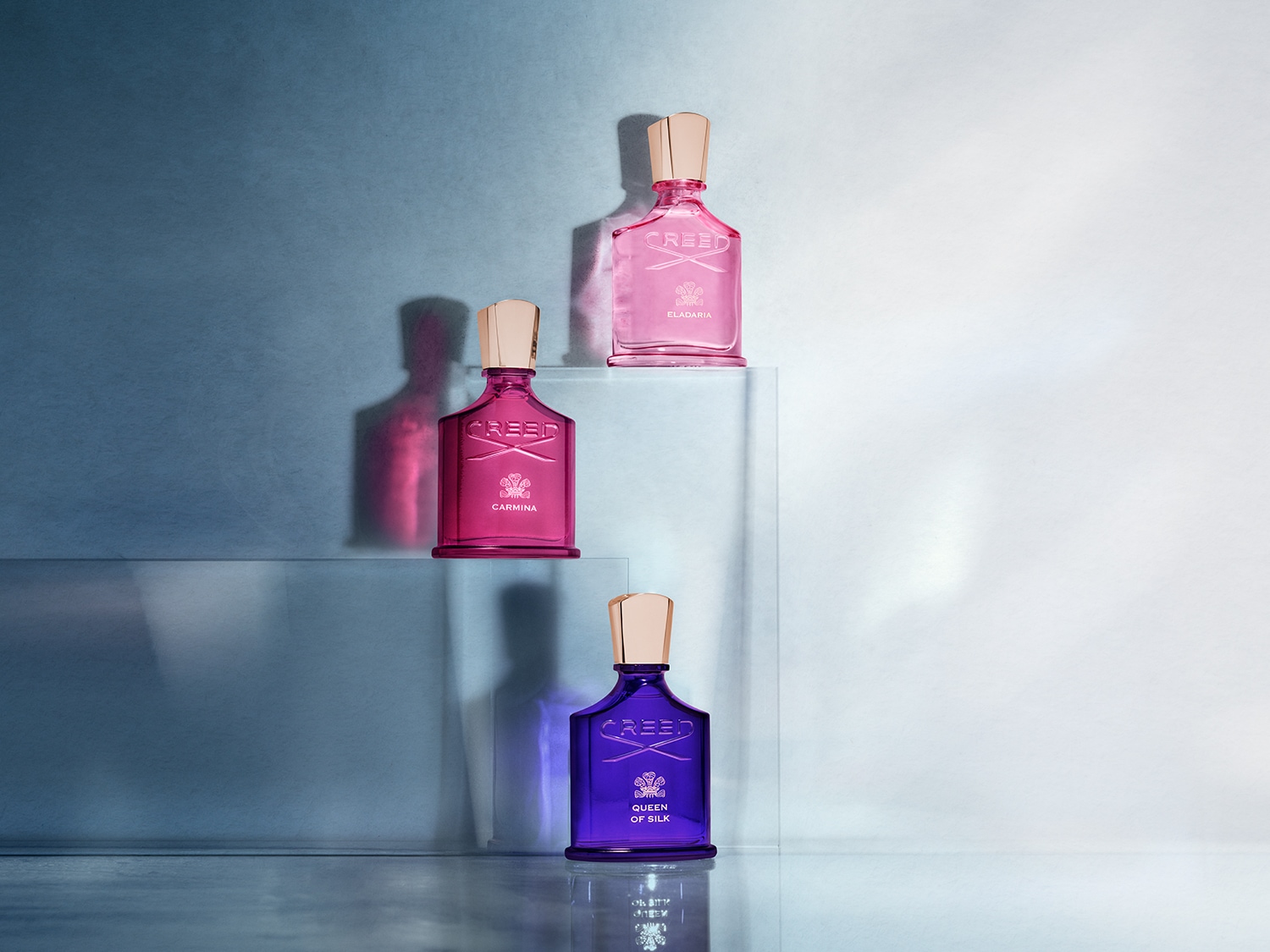 Drei Creed-Parfümflaschen in Rosa-, Pink- und Lilatönen stehen auf transparenten Sockeln und zeigen die Eleganz und Vielfalt der Düfte.