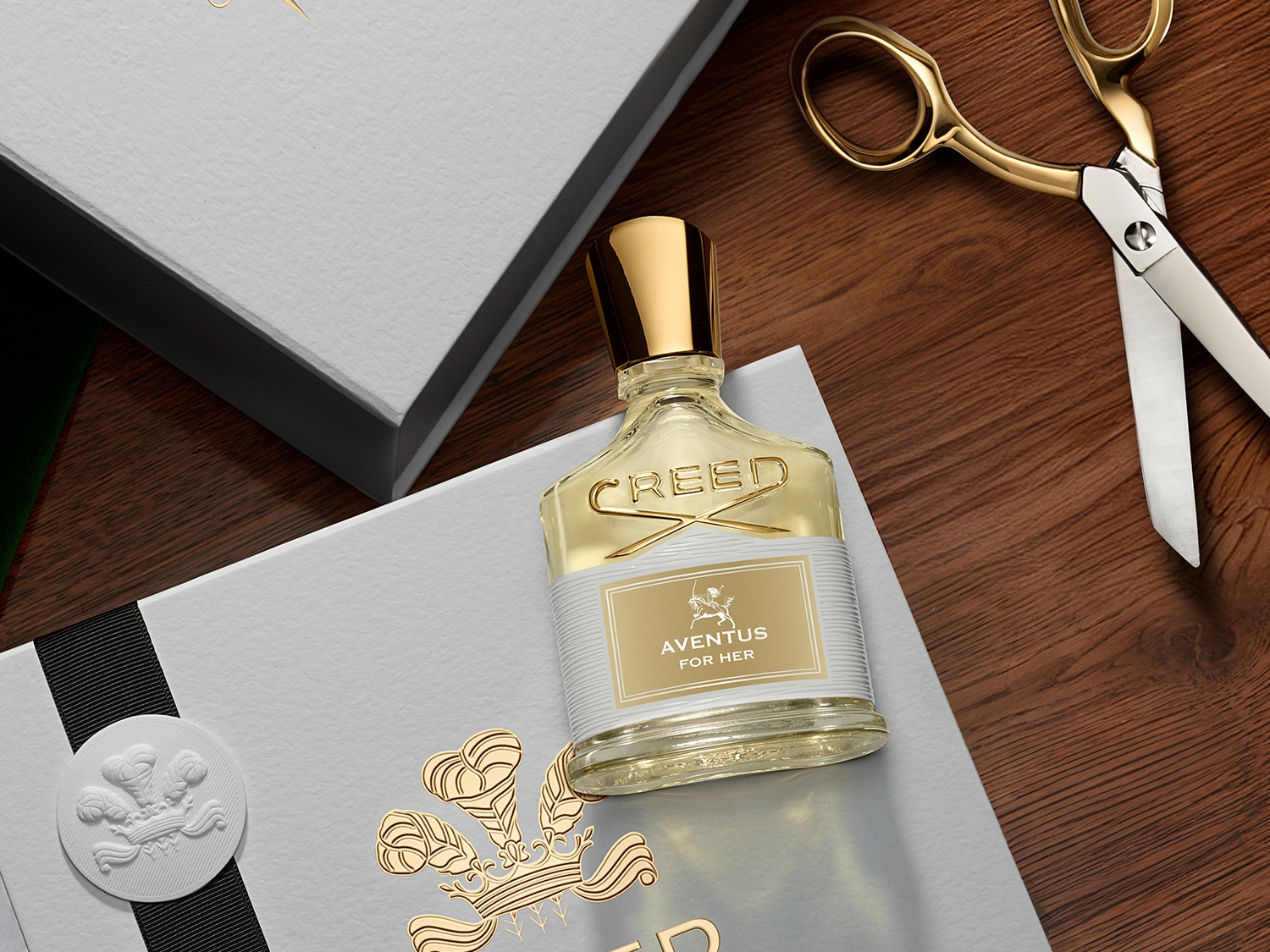 Ein Flakon Creed Aventus for Her Eau de Parfum mit goldenem Verschluss und Etikett liegt auf einer grauen Geschenkbox mit goldener Krone, daneben eine goldene Schere.