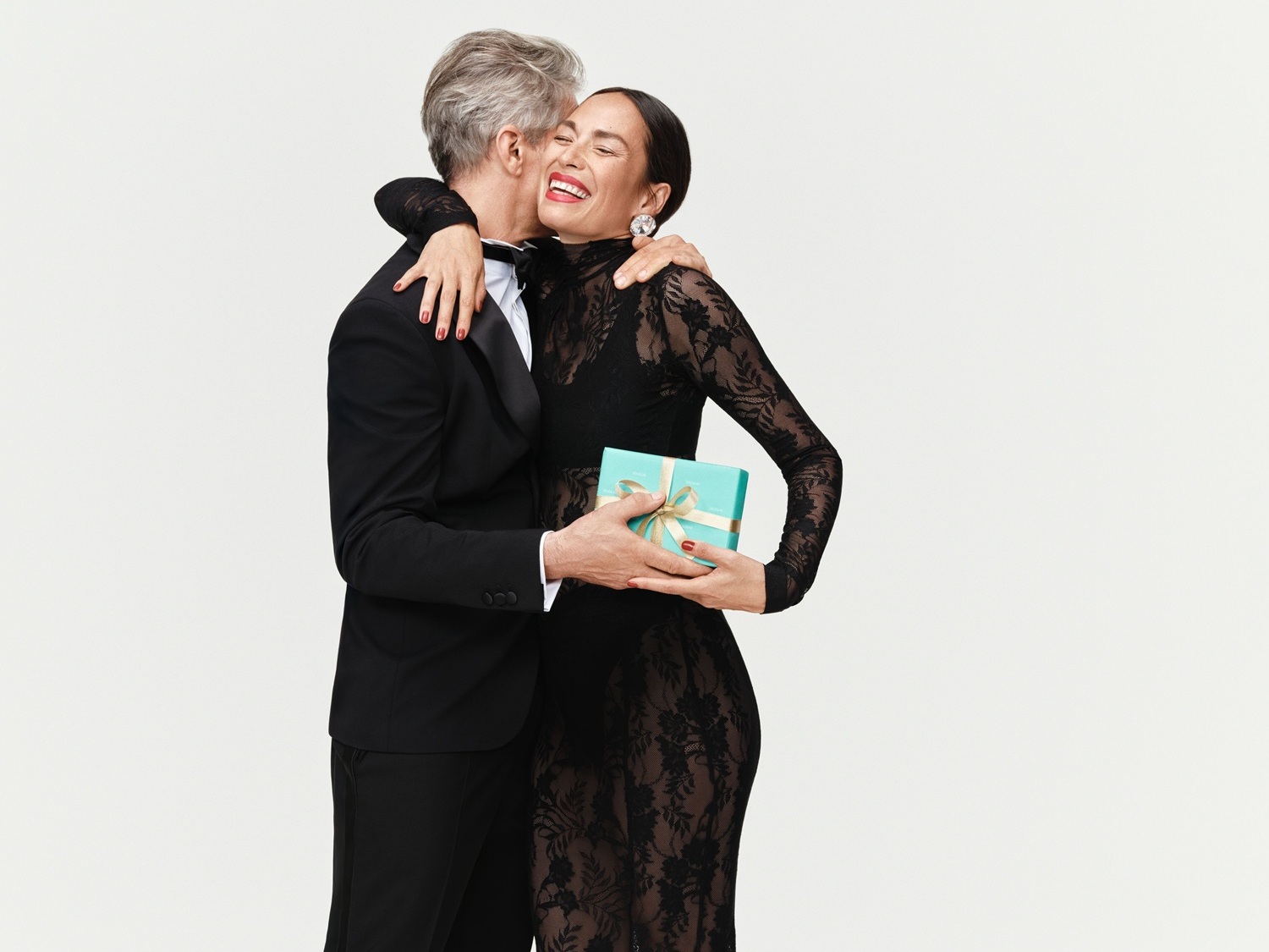 Ein Mann im Smoking umarmt eine Frau im schwarzen Spitzenkleid, die eine hellblaue Schachtel mit goldener Schleife hält.