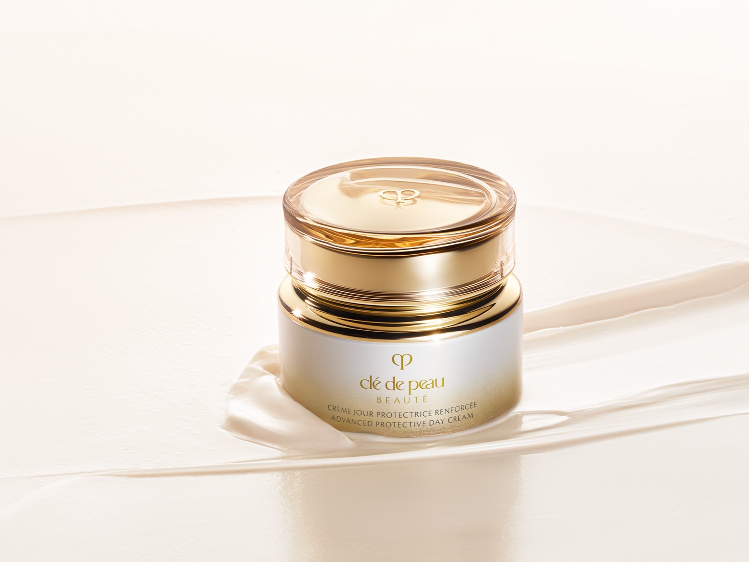 Nahaufnahme einer goldenen und weißen Dose „Clé de Peau Beauté Advanced Protective Day Cream“ auf einem cremefarbenen, glänzenden Untergrund.