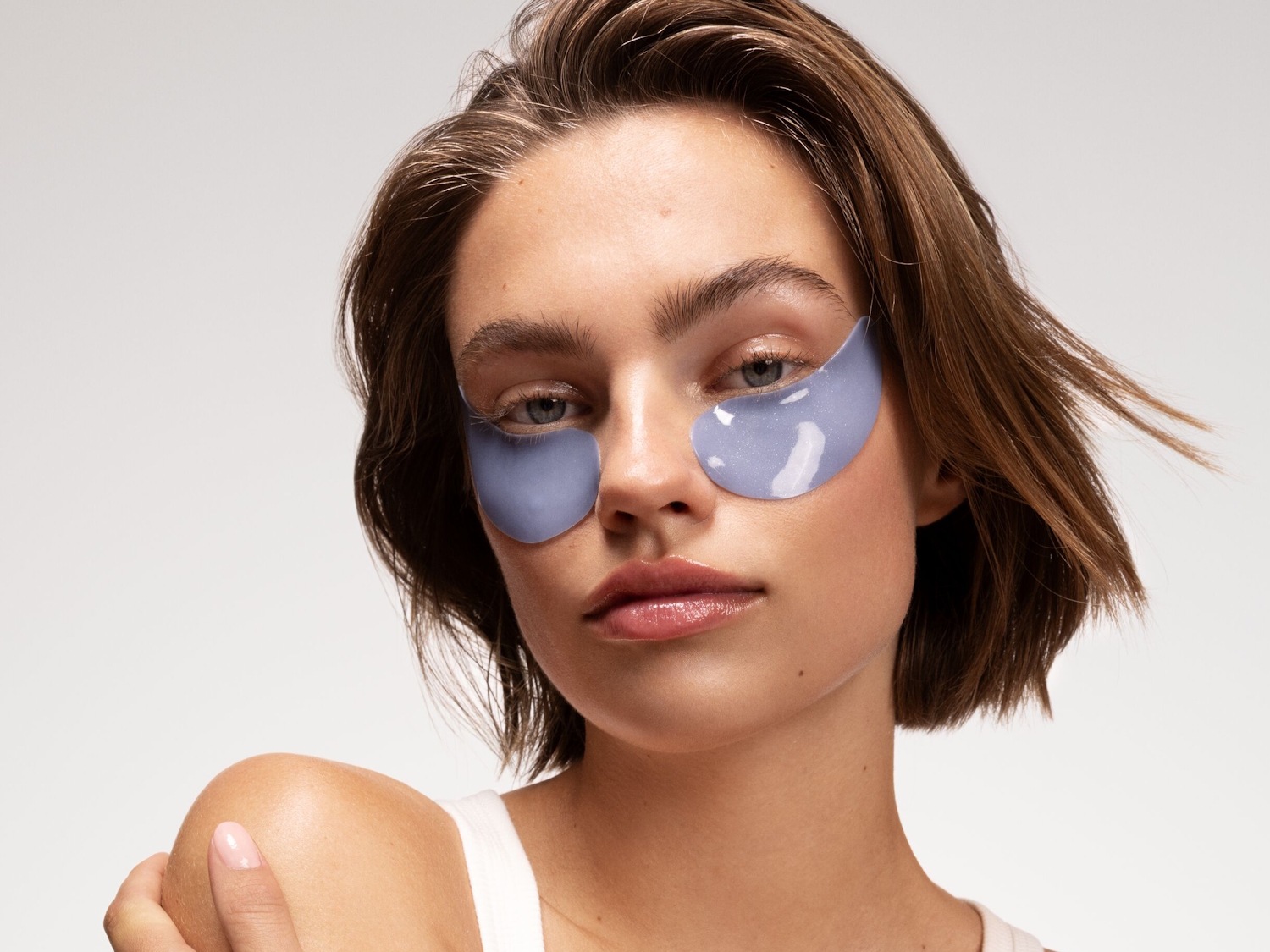 Jonge vrouw met korte bruine haren en blauwe oogmaskers onder haar ogen, voor een frisse en stralende blik.