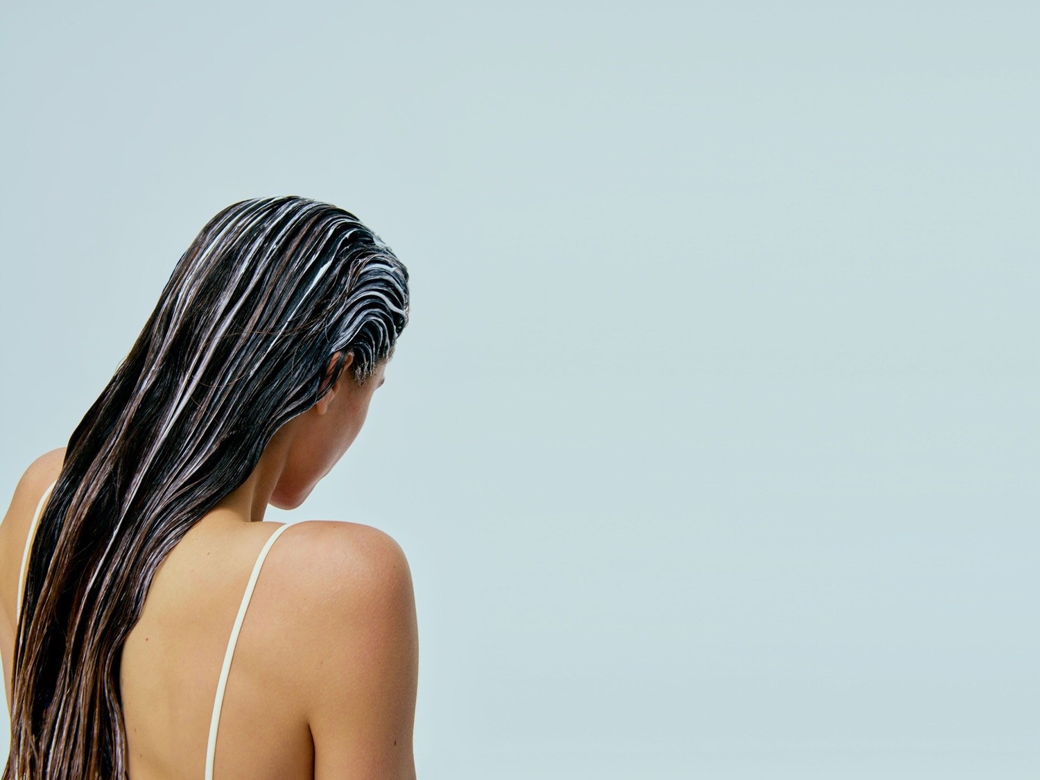 Een vrouw van achteren gezien met lang, donker haar dat bedekt is met een witte crème, mogelijk een haarmasker, tegen een lichtblauwe achtergrond.