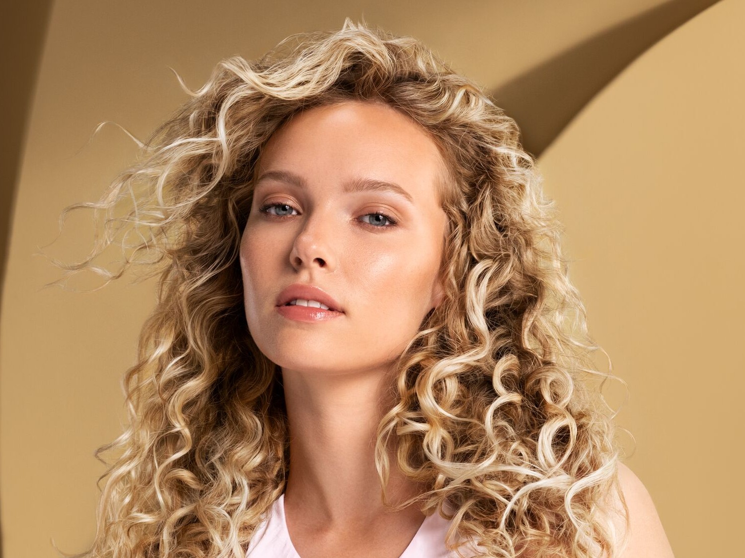 Portret van een vrouw met blond krullend haar en blauwe ogen tegen een beige achtergrond.