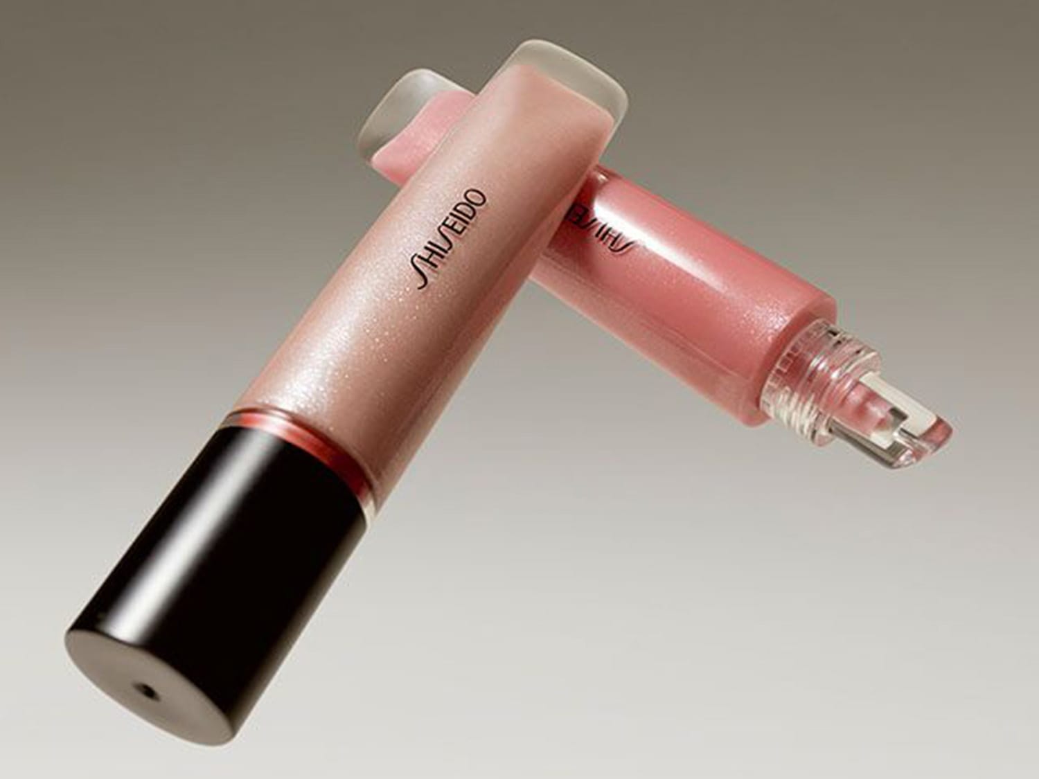 Zwei Shiseido Lipgloss-Tuben liegen übereinander, eine in einem schimmernden Nude-Ton mit schwarzem Deckel und die andere in einem zarten Rosa mit transparentem Applikator.