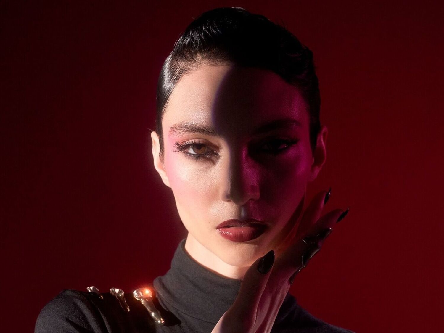 Portrait d'une femme avec un maquillage audacieux, des ombres roses, un eye-liner noir et des lèvres rouge foncé, portant un col roulé noir orné de bijoux dorés, le tout sur un fond rouge profond.
