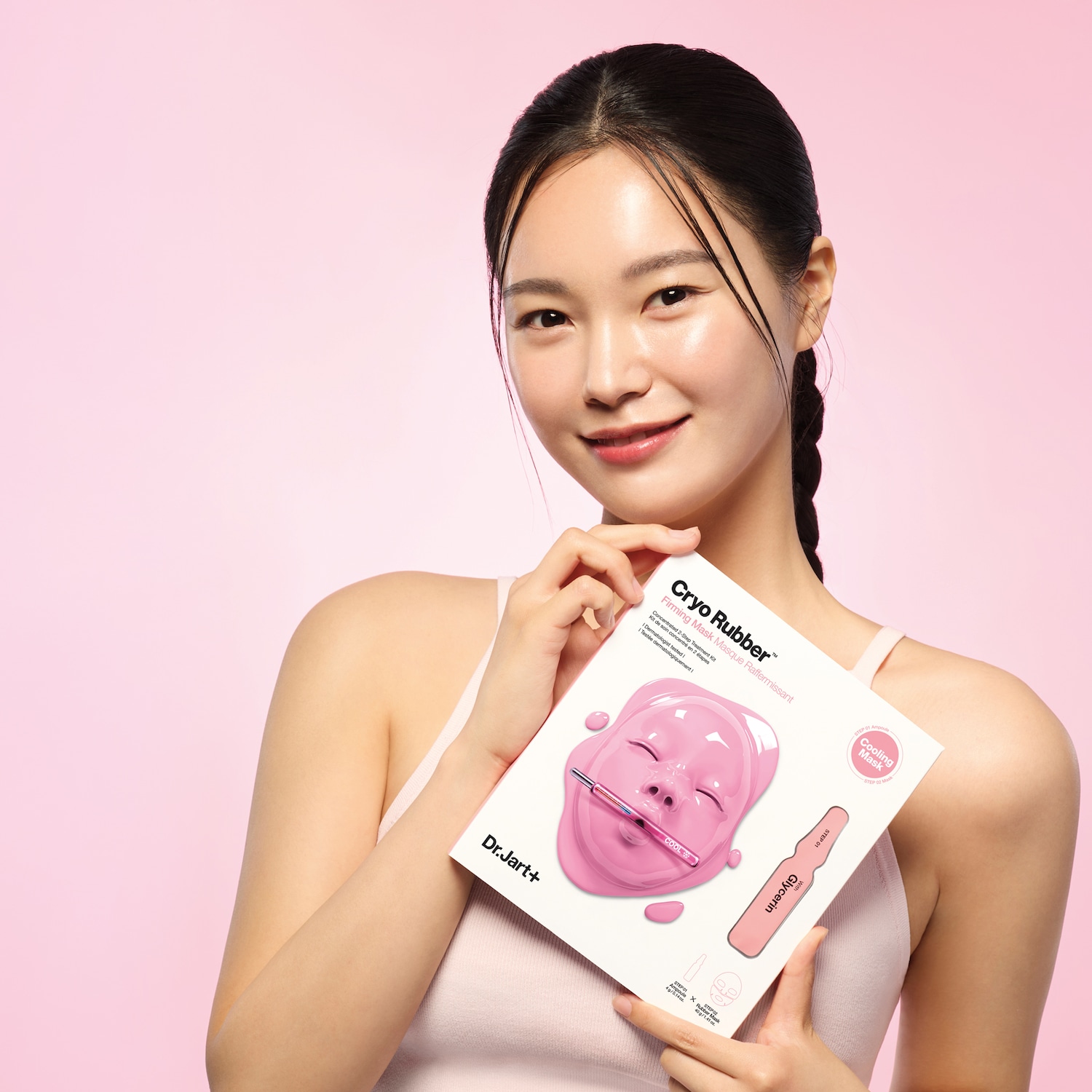 Een jonge vrouw toont een Dr.Jart+ Cryo Rubber verstevigend masker, een 2-staps behandeling met glycerine, tegen een roze achtergrond.