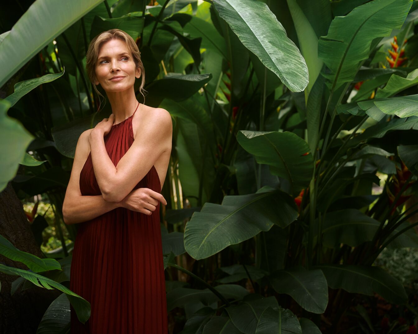 Eine Frau mit blonden Haaren, die ein ärmelloses, plissiertes, weinrotes Kleid trägt, steht inmitten üppiger grüner tropischer Pflanzen.