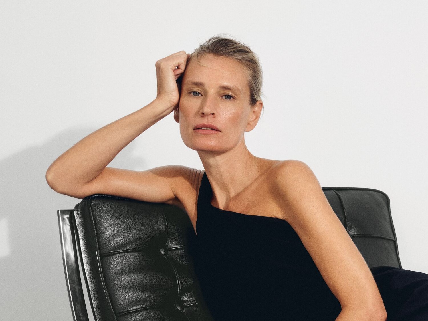 Eine Frau mit blonden, zurückgekämmten Haaren sitzt in einem schwarzen, trägerlosen Oberteil auf einem schwarzen Ledersessel und stützt ihren Kopf auf ihre Hand.