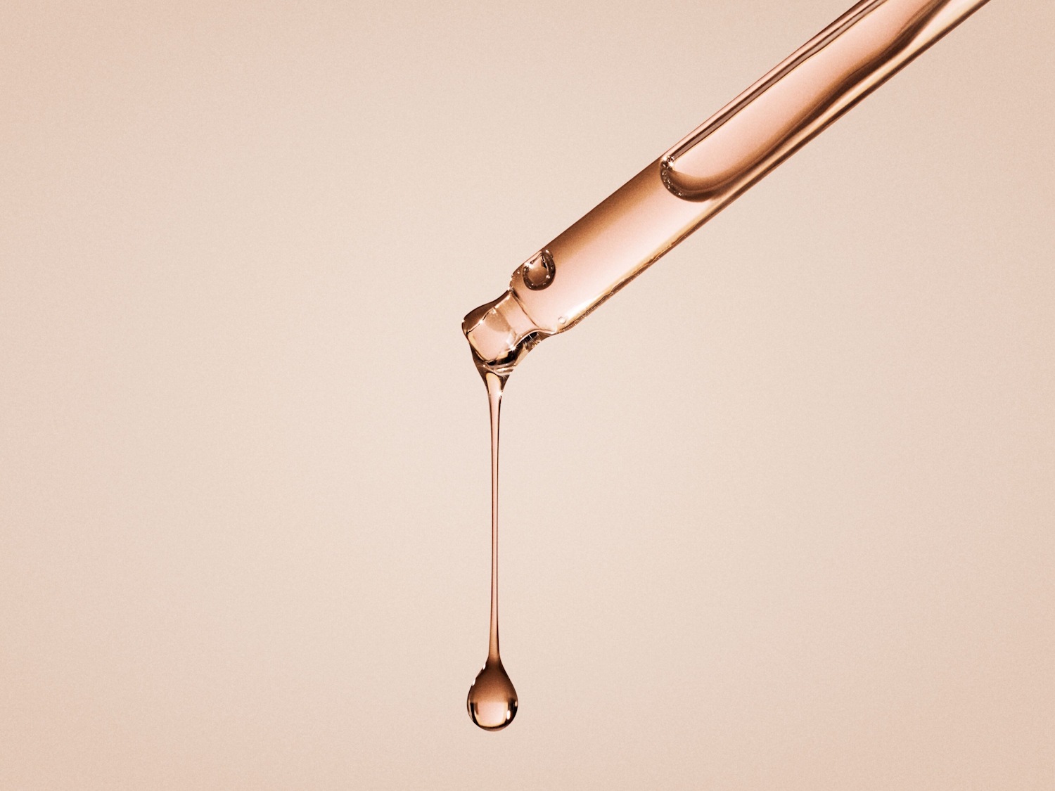 Close-up van een glazen pipet met een druppel roze serum die eraan hangt, tegen een zachtroze achtergrond.