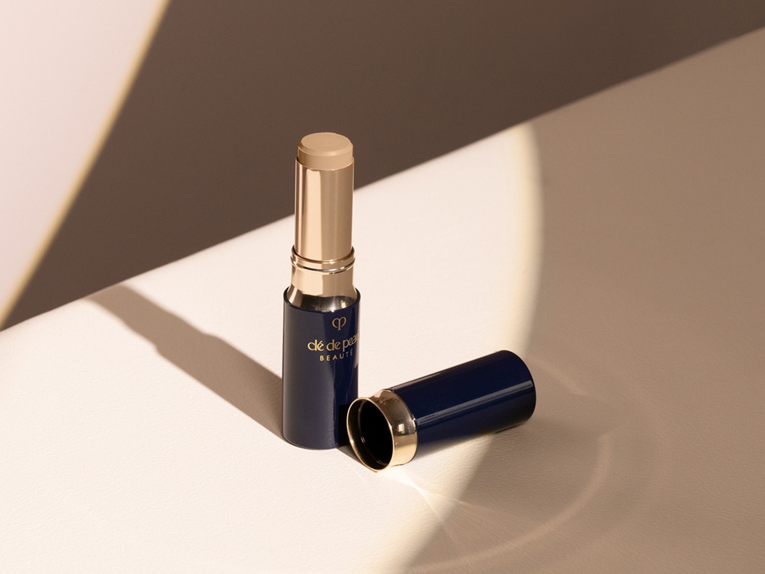 Voici un stick de fond de teint Clé de Peau Beauté, son emballage est bleu marine et or, le capuchon est retiré pour révéler le stick beige.