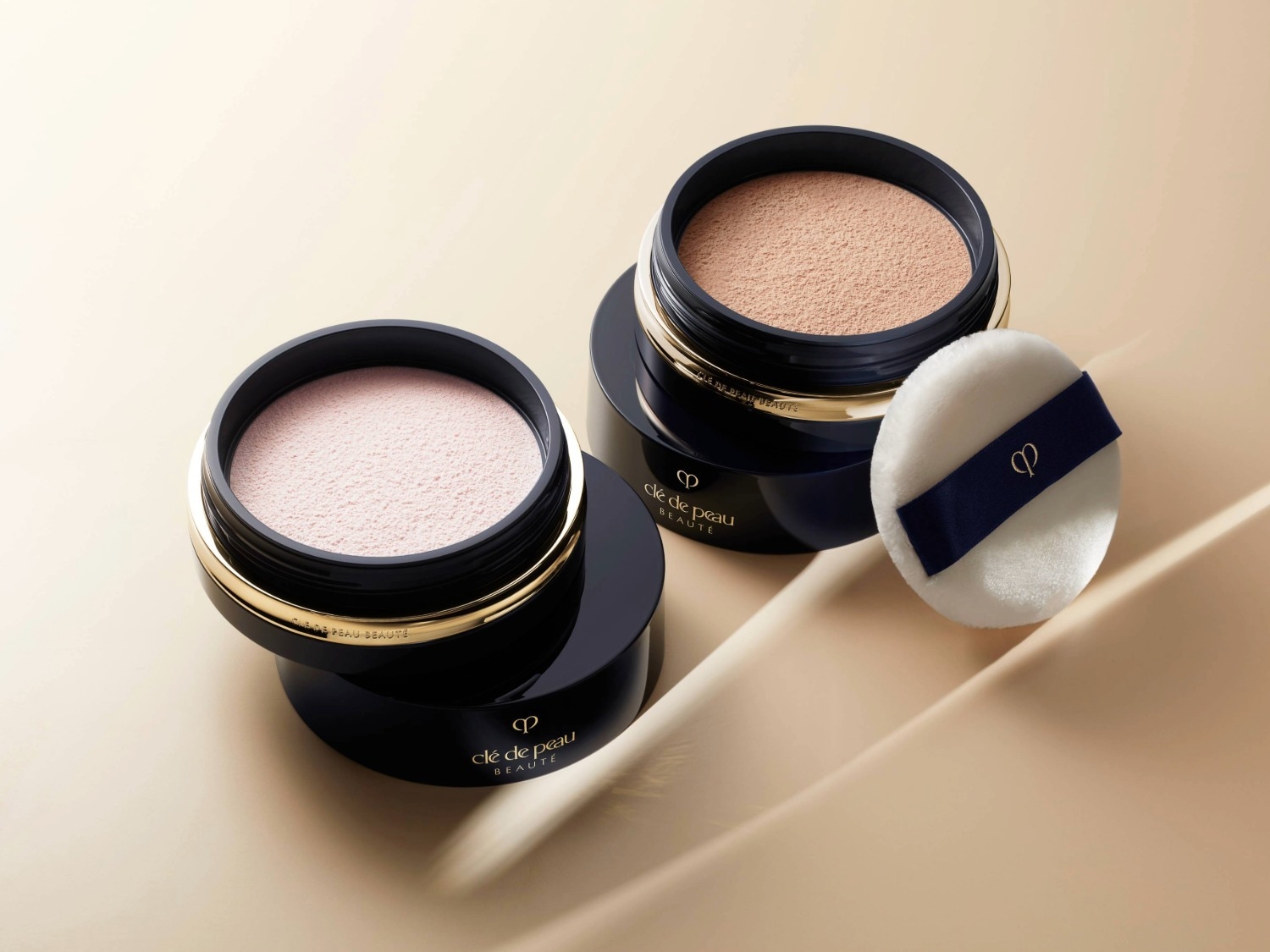 Voici deux boîtiers de poudre compacte Clé de Peau Beauté, l'un avec une poudre rose pâle et l'autre avec une poudre beige, accompagnés d'une houppette blanche ornée d'un ruban bleu marine.
