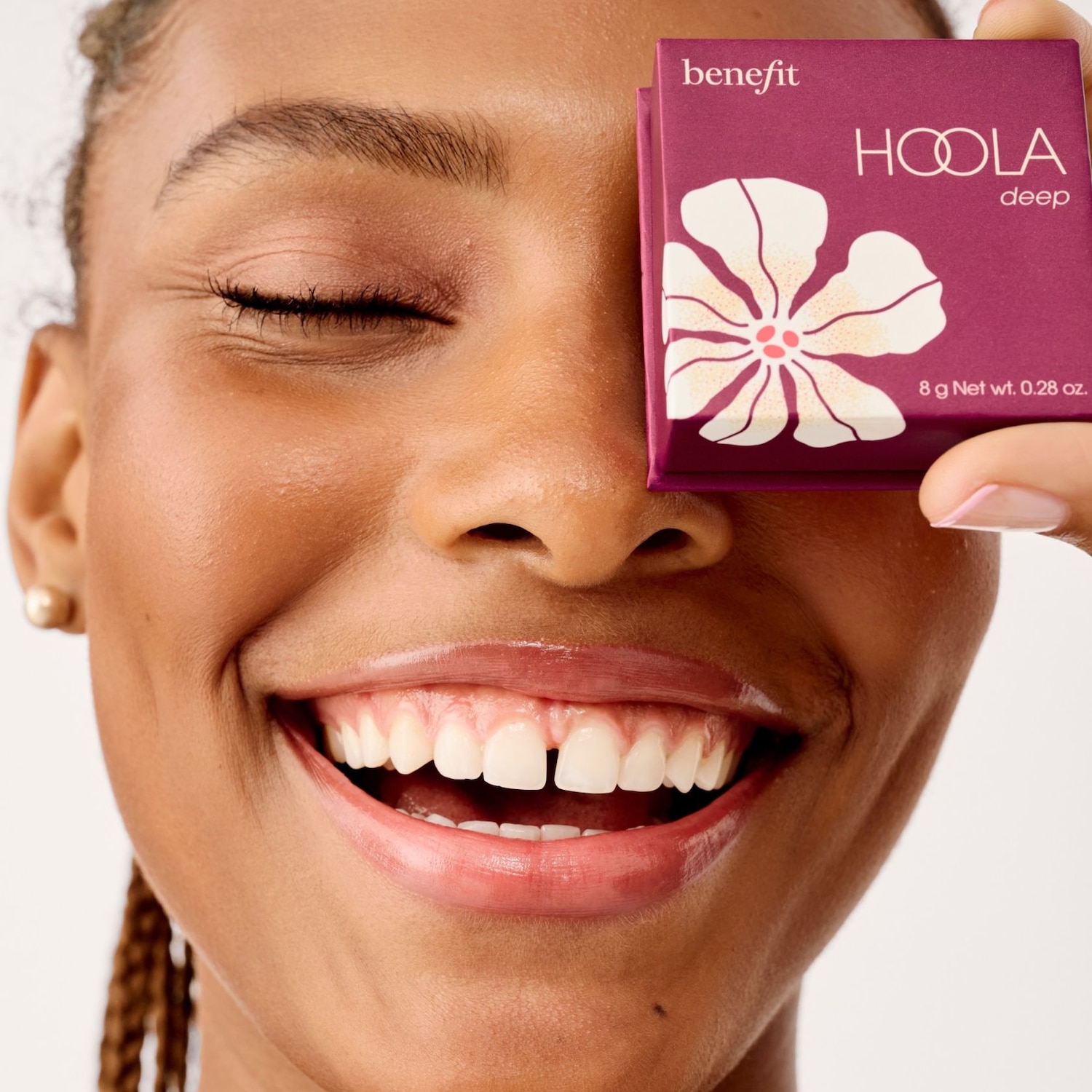 Een lachende vrouw houdt een Benefit Hoola Deep bronzer vast, de verpakking is paars met een witte bloem.