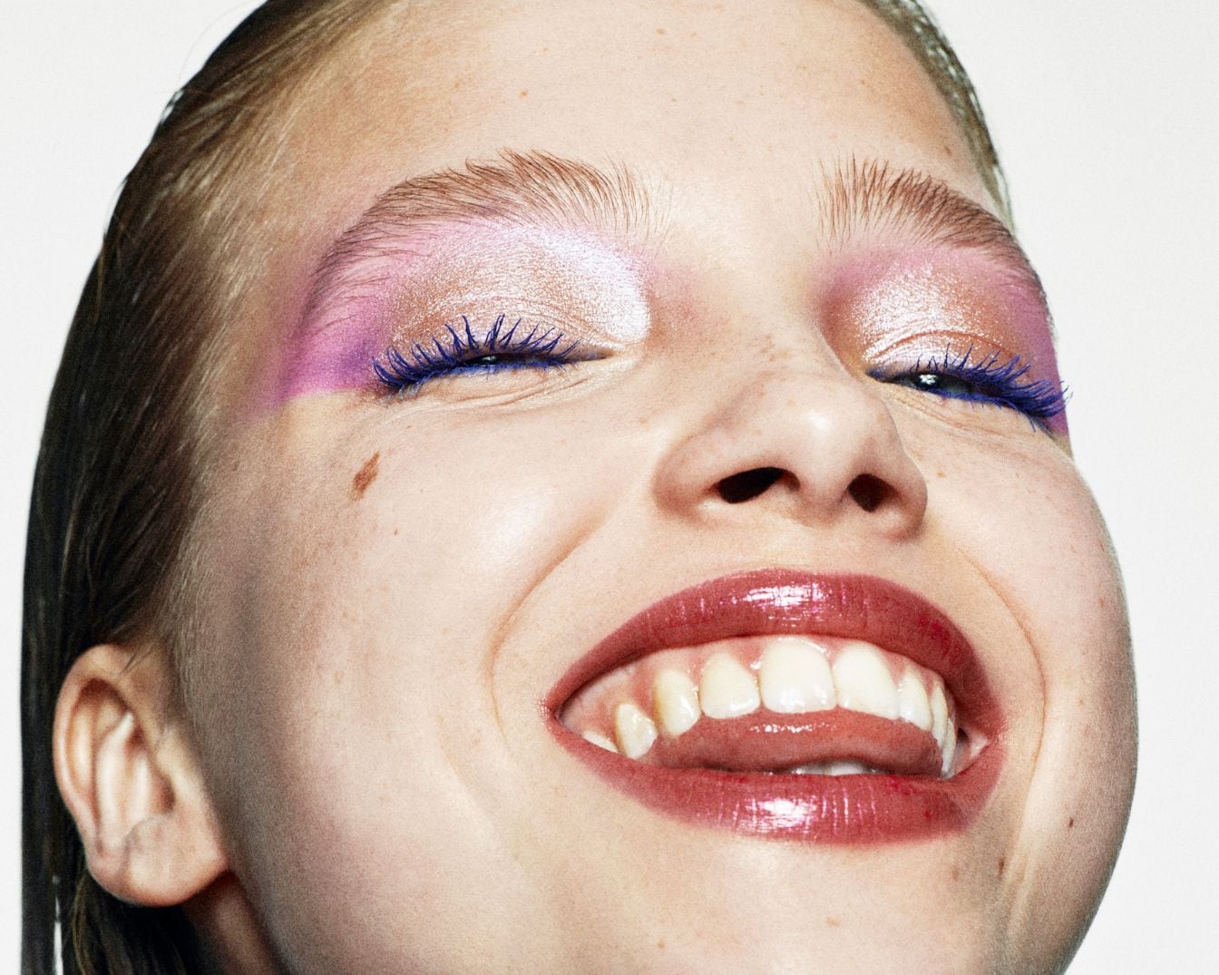 Jonge vrouw lacht met roze oogschaduw, blauwe mascara en glanzende lippenstift, waardoor een gedurfde en speelse make-uplook ontstaat.