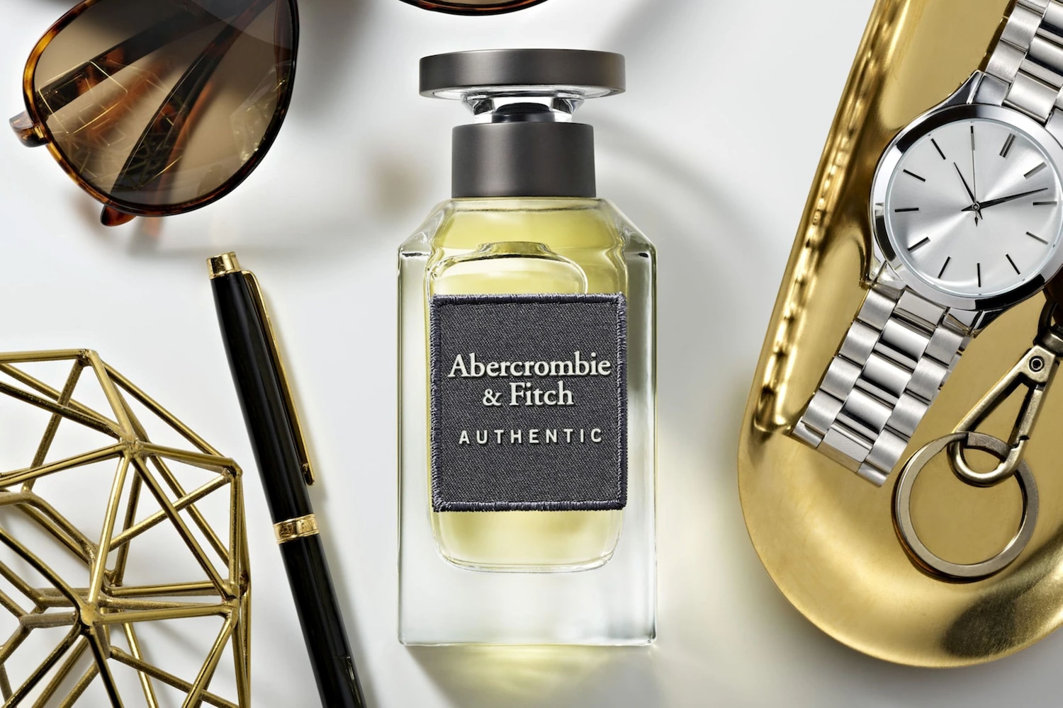 Das Abercrombie & Fitch Authentic Eau de Toilette präsentiert sich in einem klaren Glasflakon mit einem grauen Etikett, umgeben von stilvollen Accessoires wie einer Uhr und einer Sonnenbrille.