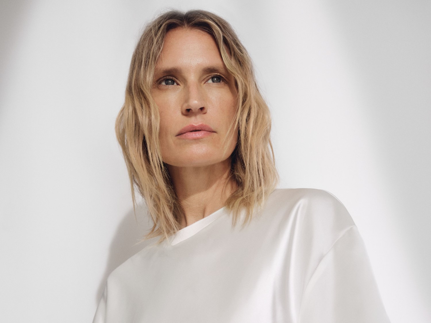 Una mujer rubia con una blusa blanca de seda mira hacia arriba con una expresión serena, resaltando la elegancia y la sencillez en un fondo claro.