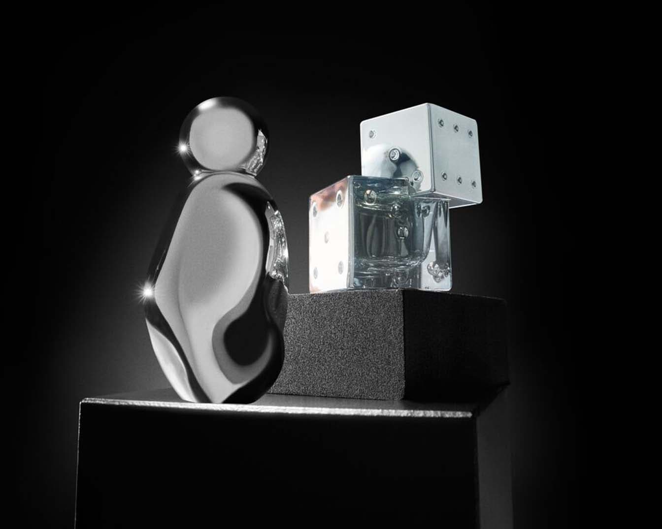 Ezüstös, gömbölyded üvegcsében parfüm, mellette kocka alakú, ezüstös kupakú üvegcsék állnak egy fekete alapon.