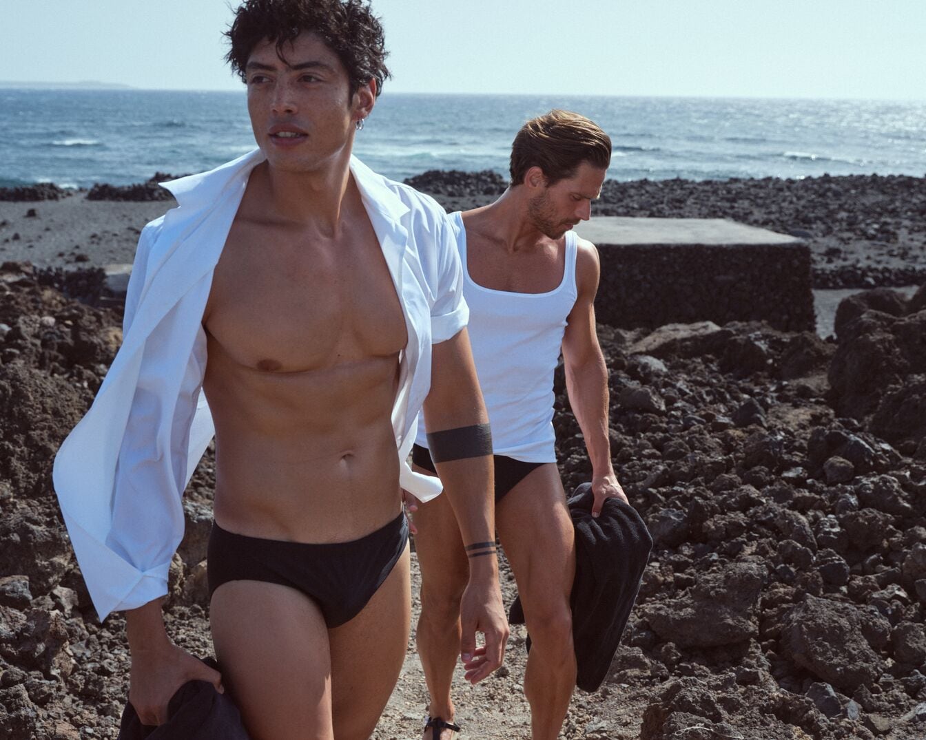 Zwei Männer in Badehosen und weißen Oberteilen spazieren an einem felsigen Strand entlang, das Meer im Hintergrund.