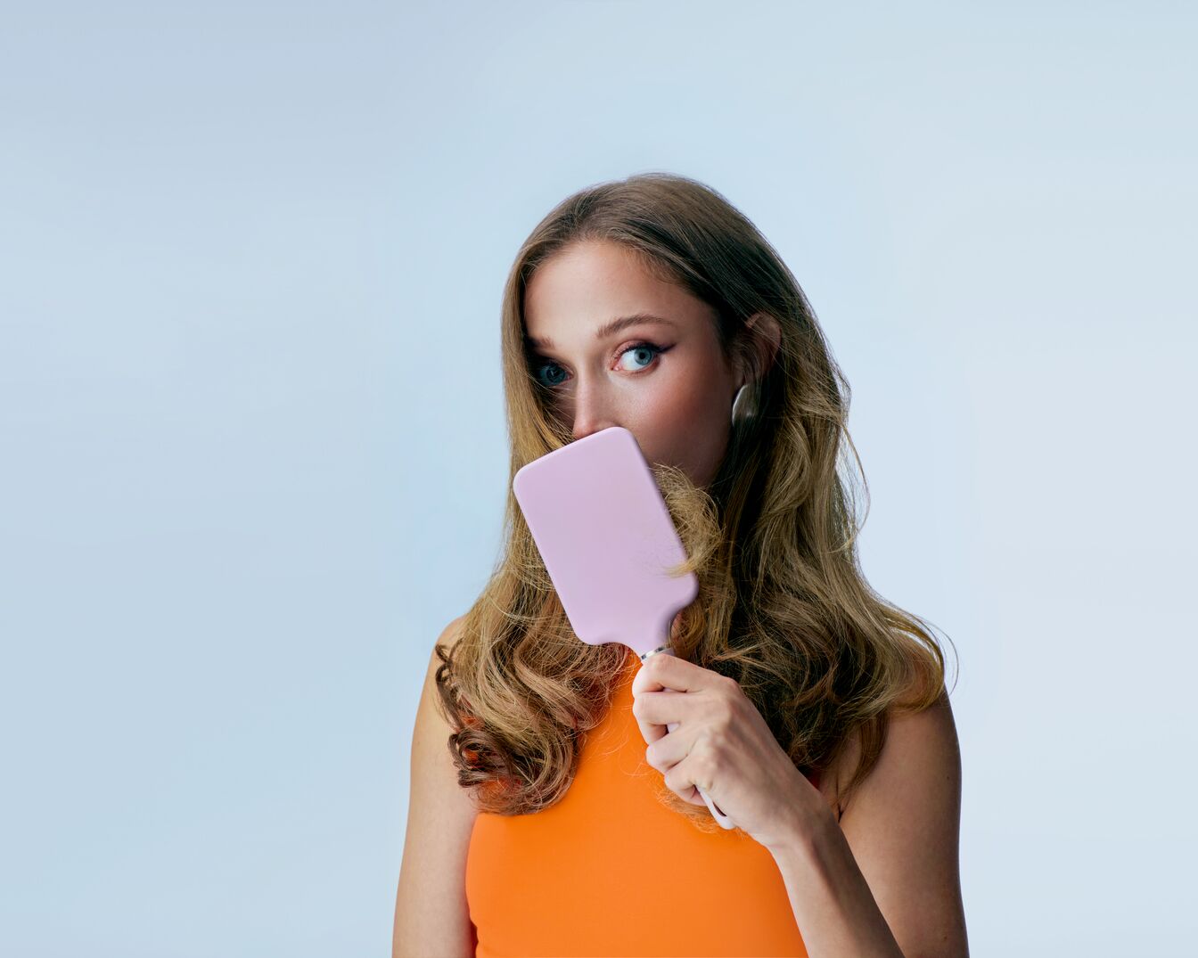 Eine Frau mit gewelltem, braunem Haar, die ein orangefarbenes Top trägt und ihr Gesicht mit einer hellrosa Haarbürste teilweise verdeckt.