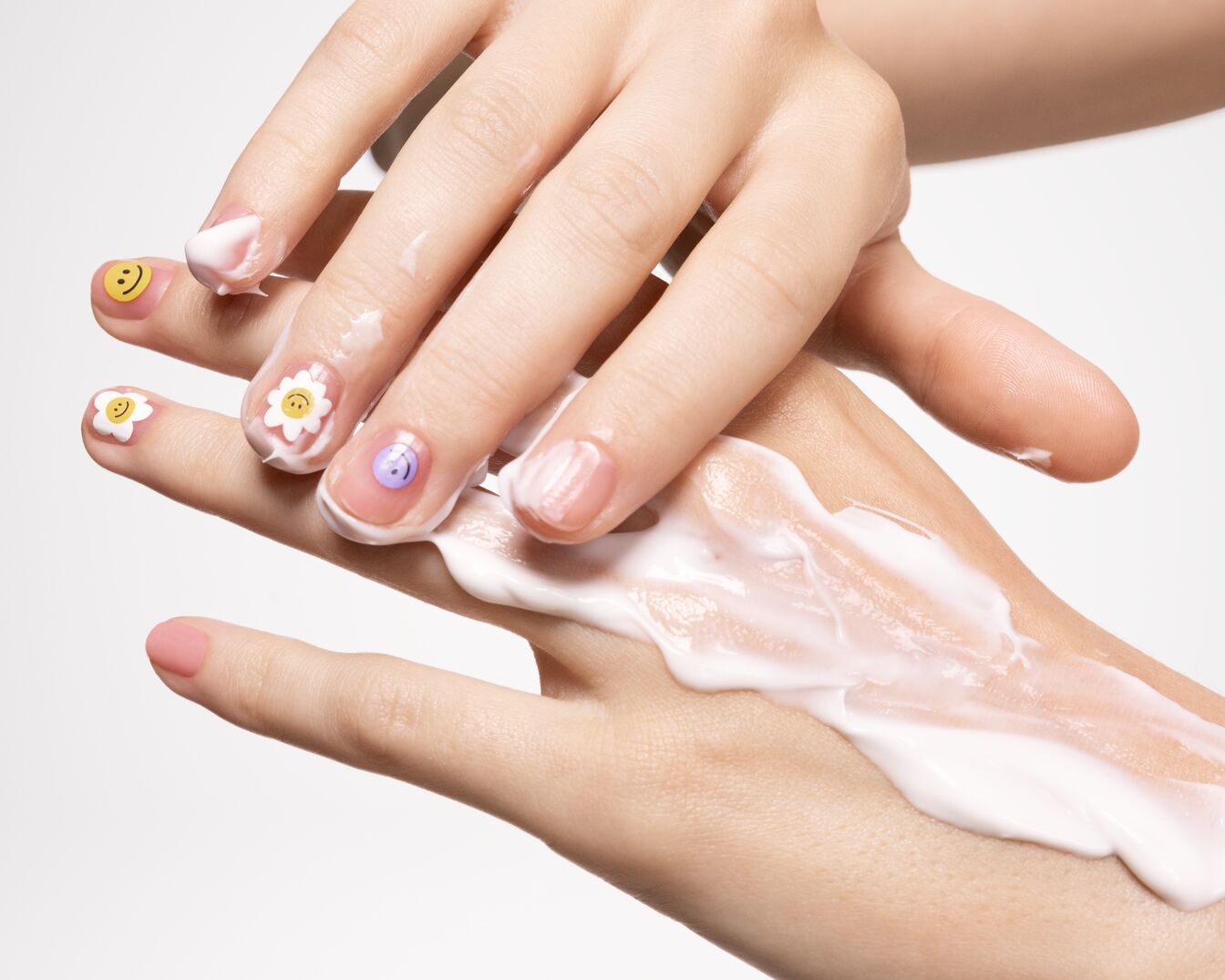 Une personne applique une crème blanche et épaisse sur le dos de sa main, ses ongles sont décorés de motifs floraux et de smileys.