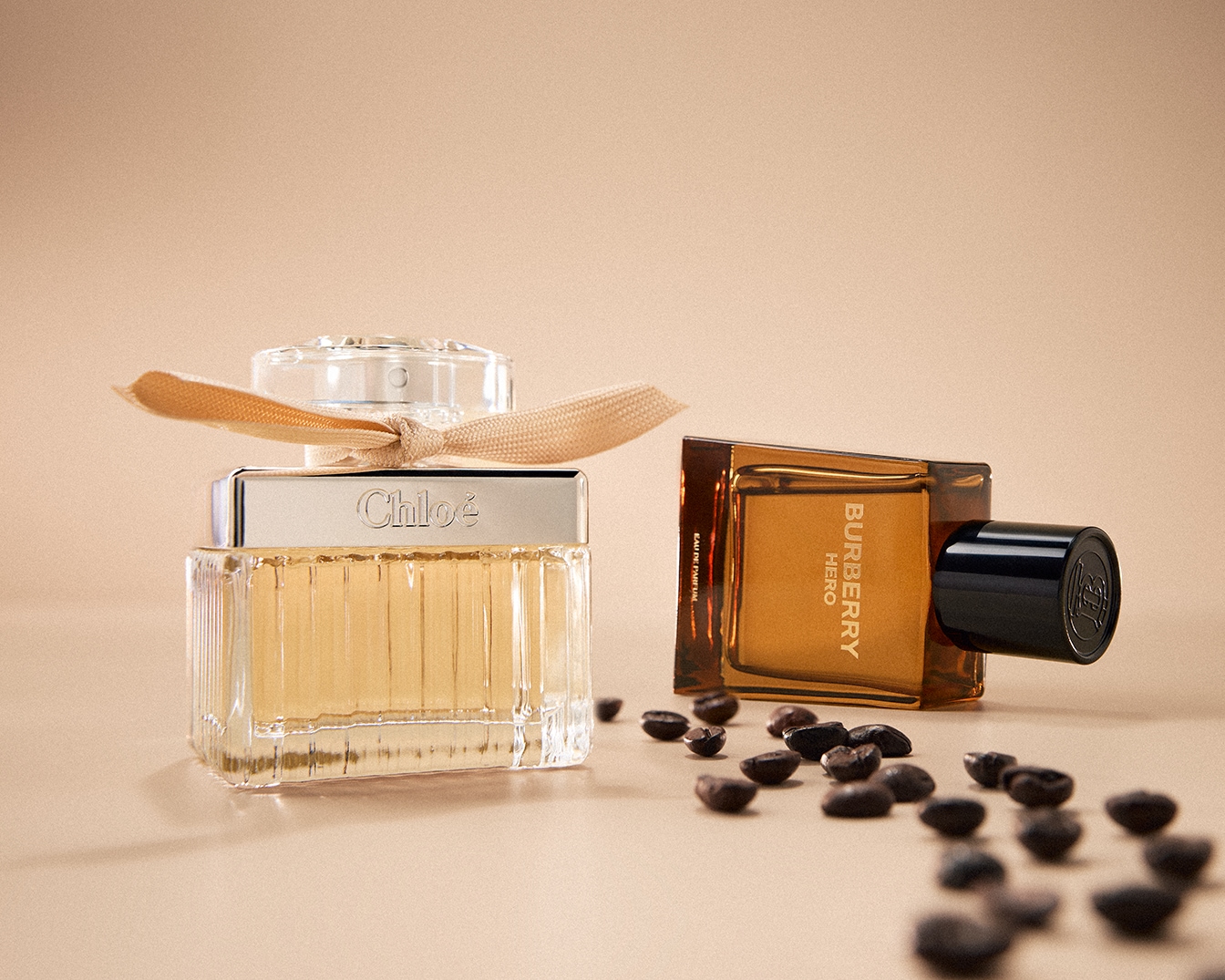 Šalia išdėlioti kavos pupelės, o fone matomi du kvepalų buteliukai: vienas – „Chloé“ su kaspinu, kitas – „Burberry Hero“ rudame buteliuke.