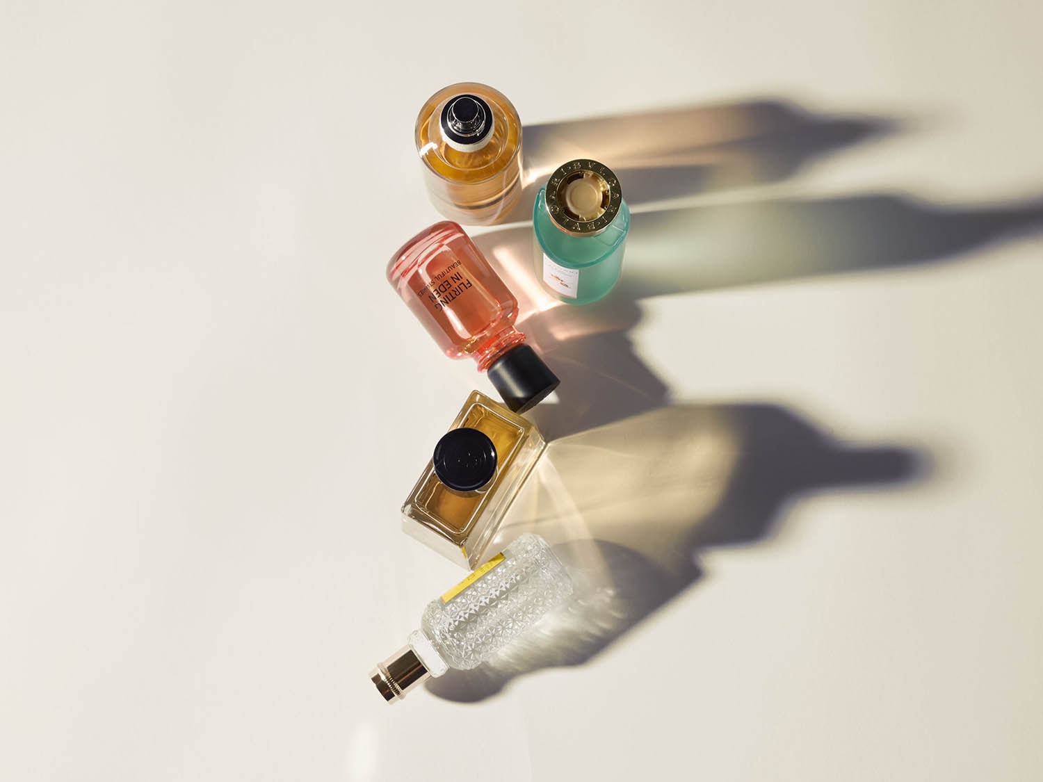 Verschiedene Parfümflaschen in Gelb-, Rosa-, Türkis- und Goldtönen liegen auf einer hellen Oberfläche und werfen lange Schatten.