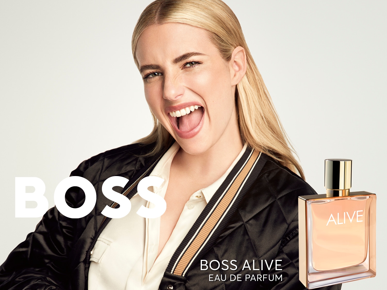 Sonriente, una mujer rubia presenta el perfume BOSS ALIVE Eau de Parfum en un frasco cuadrado con un jugo de color melocotón suave y un tapón dorado.