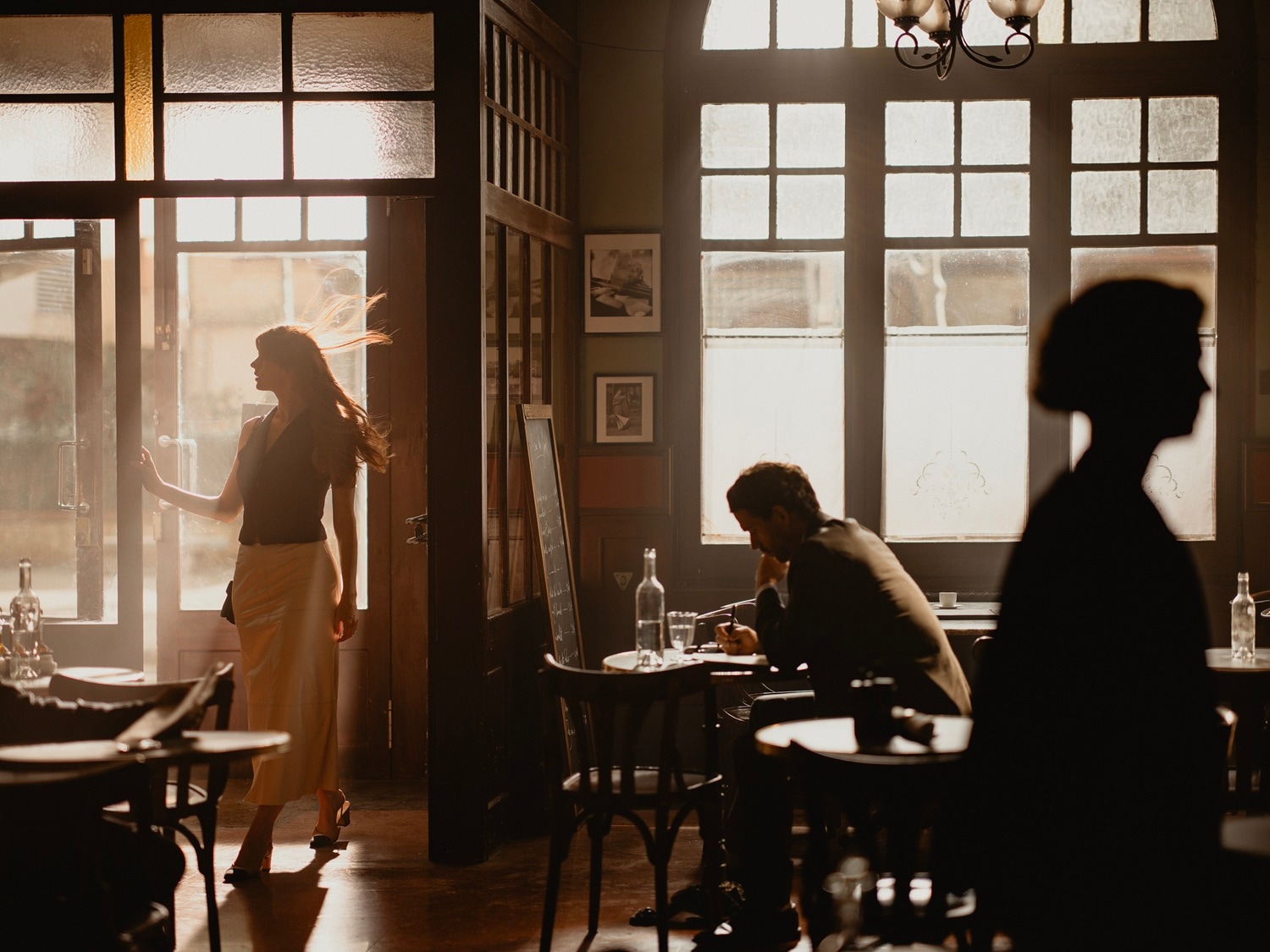 Wnętrze kawiarni oświetlone ciepłym światłem słonecznym, gdzie kobieta wchodzi przez drzwi, a mężczyzna siedzi przy stoliku.