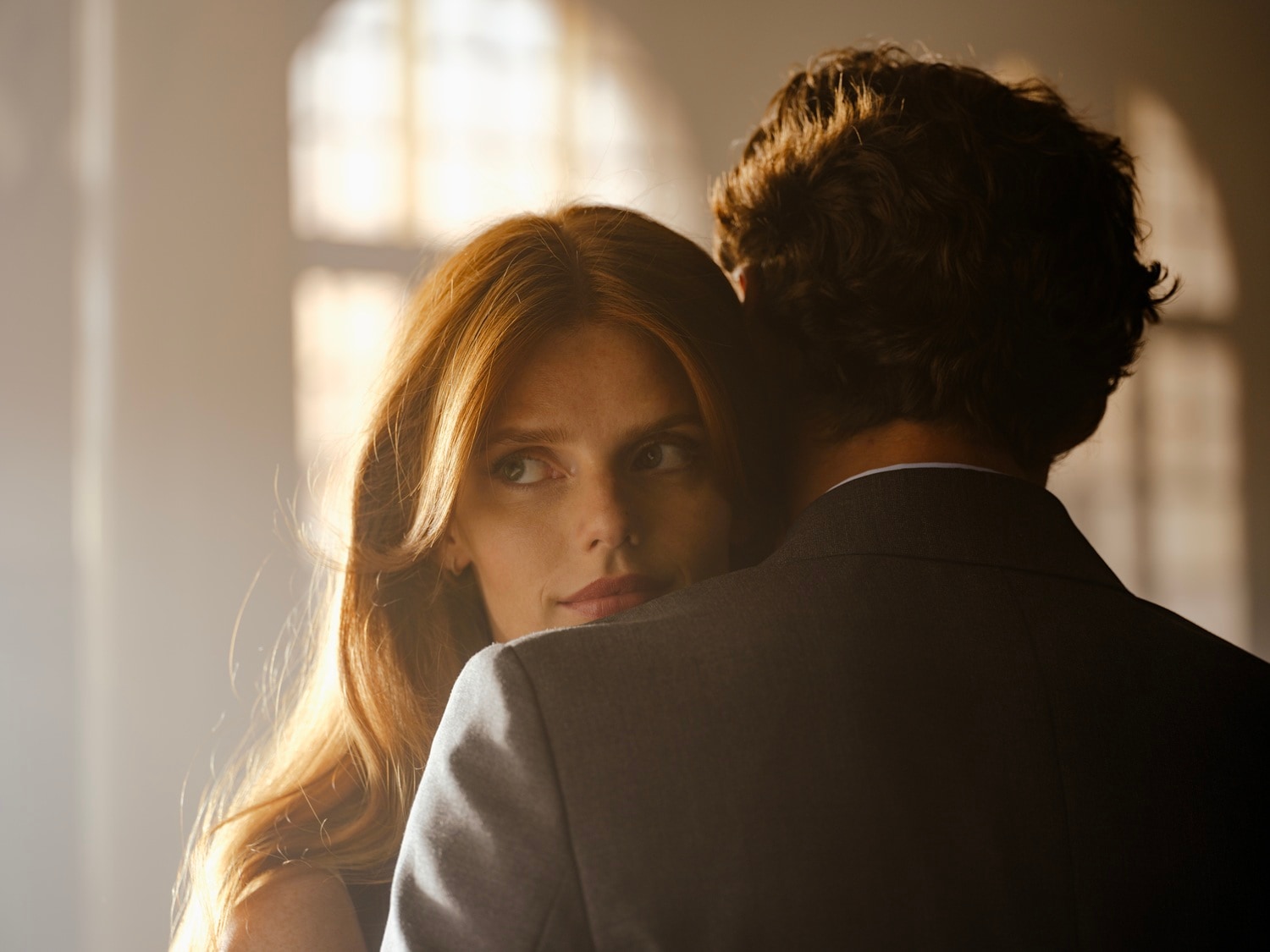 Kobieta z długimi, rudymi włosami spogląda przez ramię mężczyzny w garniturze, tworząc intymną i pełną napięcia scenę.