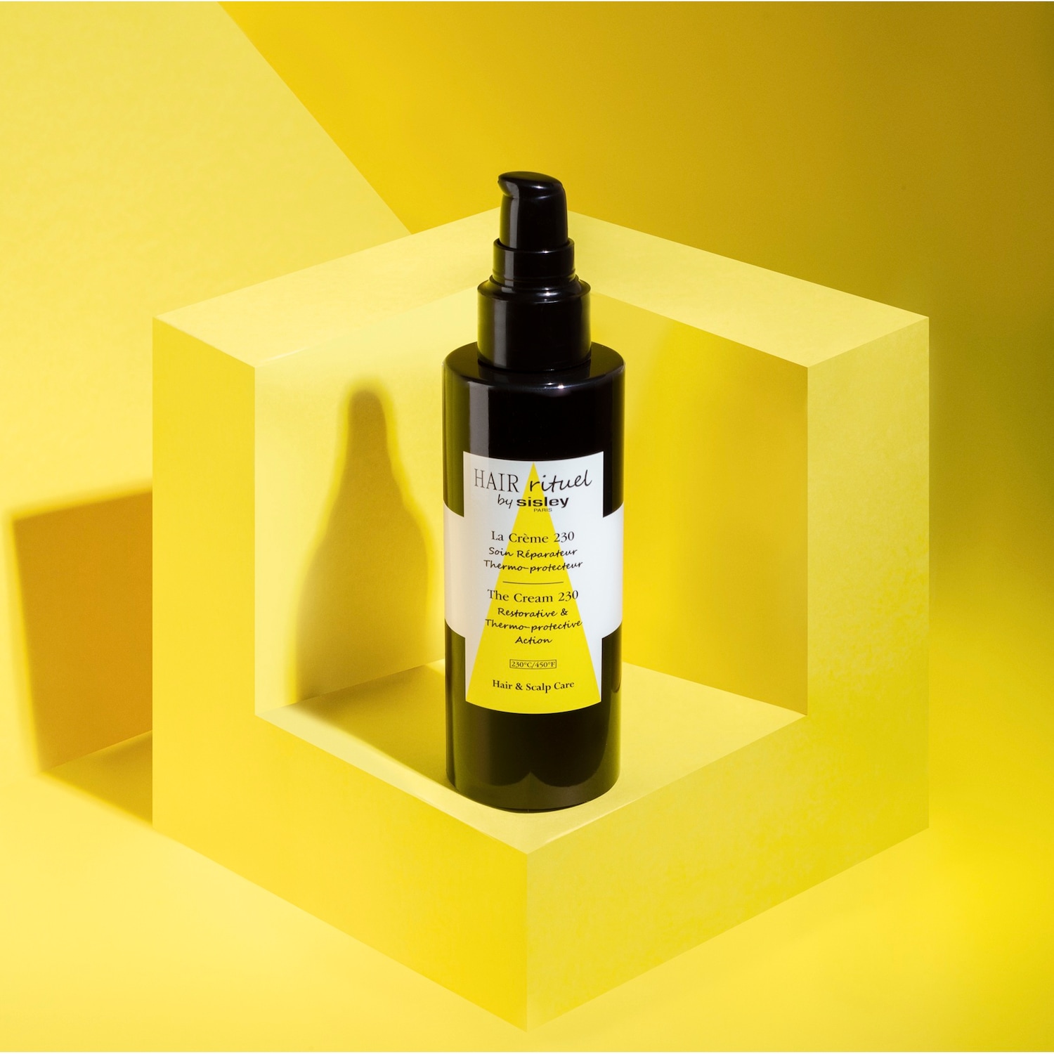 Czarna butelka Hair Rituel by Sisley La Crème 230 z pompką na żółtym tle, krem do włosów i skóry głowy o działaniu odbudowującym i termo-ochronnym.