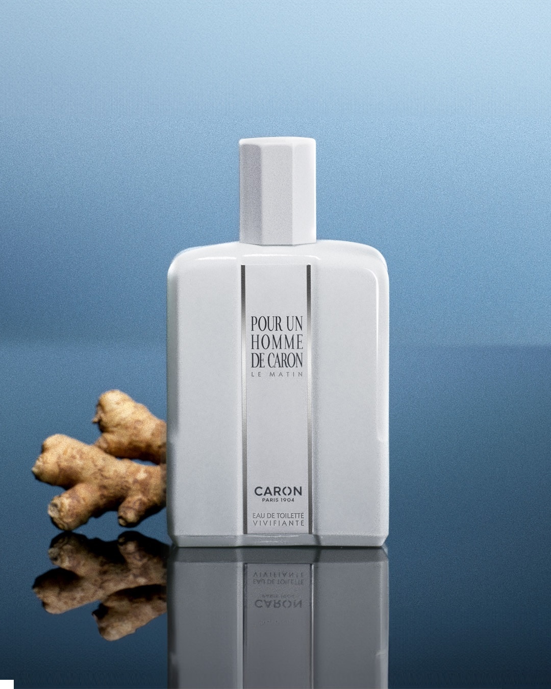 Biały flakon perfum Caron Pour Un Homme De Caron Le Matin Eau de Toilette Vivifiante z korzeniem imbiru obok, na błyszczącym tle.