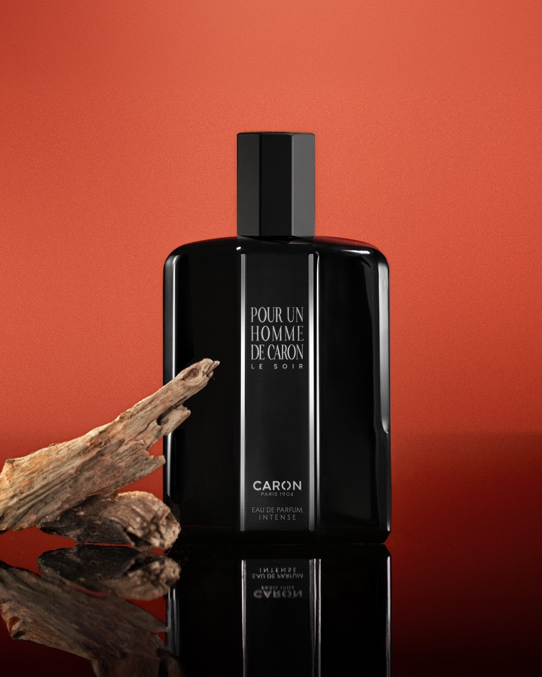 Czarny flakon perfum Caron Pour Un Homme Le Soir Eau de Parfum Intense, z drewnianymi elementami dekoracyjnymi, na błyszczącym tle.